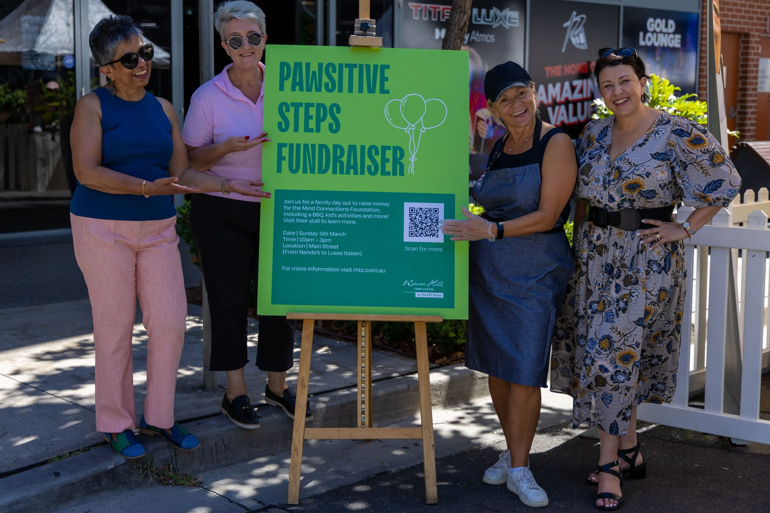 Four women standing on sidewalk, smiling at camera, pointing at a large green sign that reads 'Pawsitive Steps Fundraiser.' The sign displays event details and a QR code, with a background of shops and storefronts.