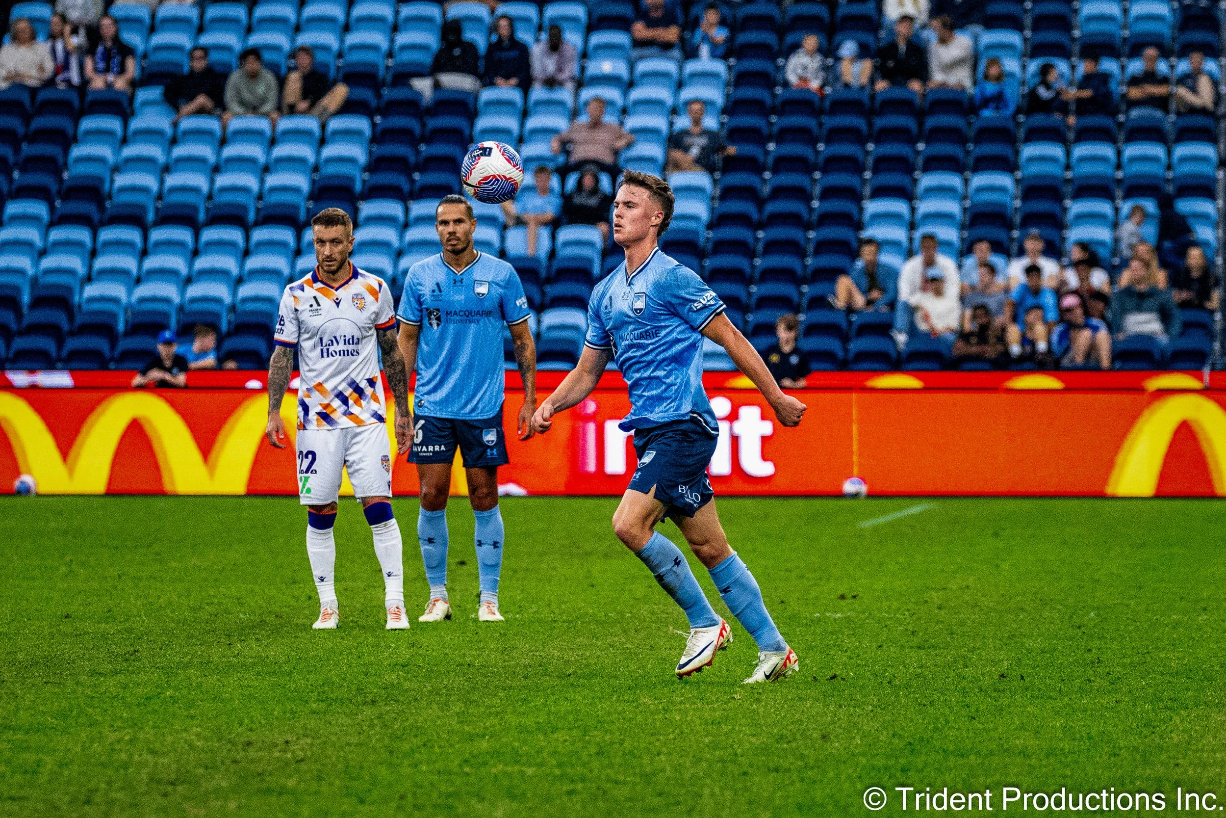 A soccer player in a light blue uniform is preparing to kick a soccer ball during a match. Two other players in white and blue uniforms are watching, with a sparse crowd in the empty blue stadium seats behind them.