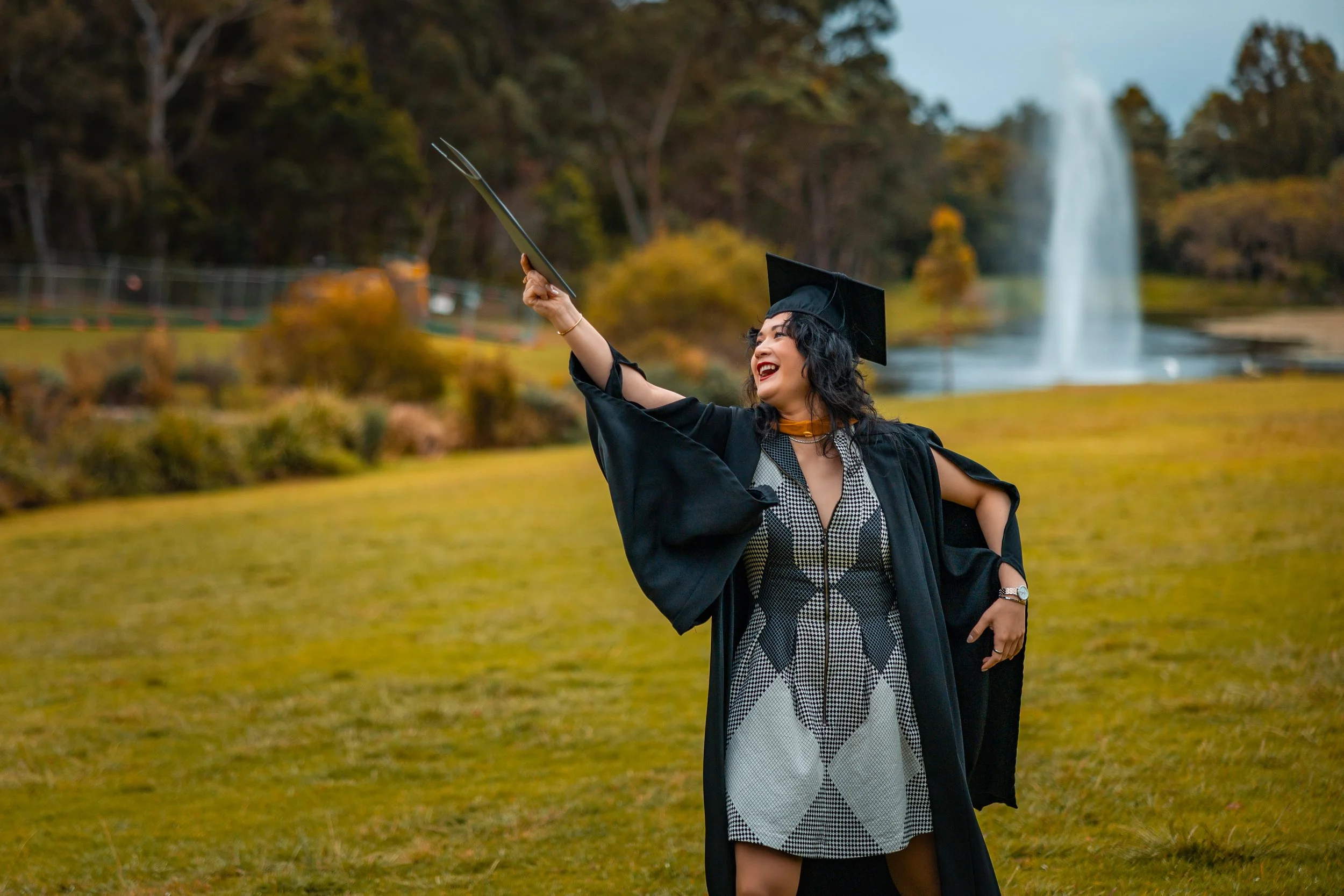 A woman in graduation robes and cap celebrating outdoors with a fountain and trees in the background.