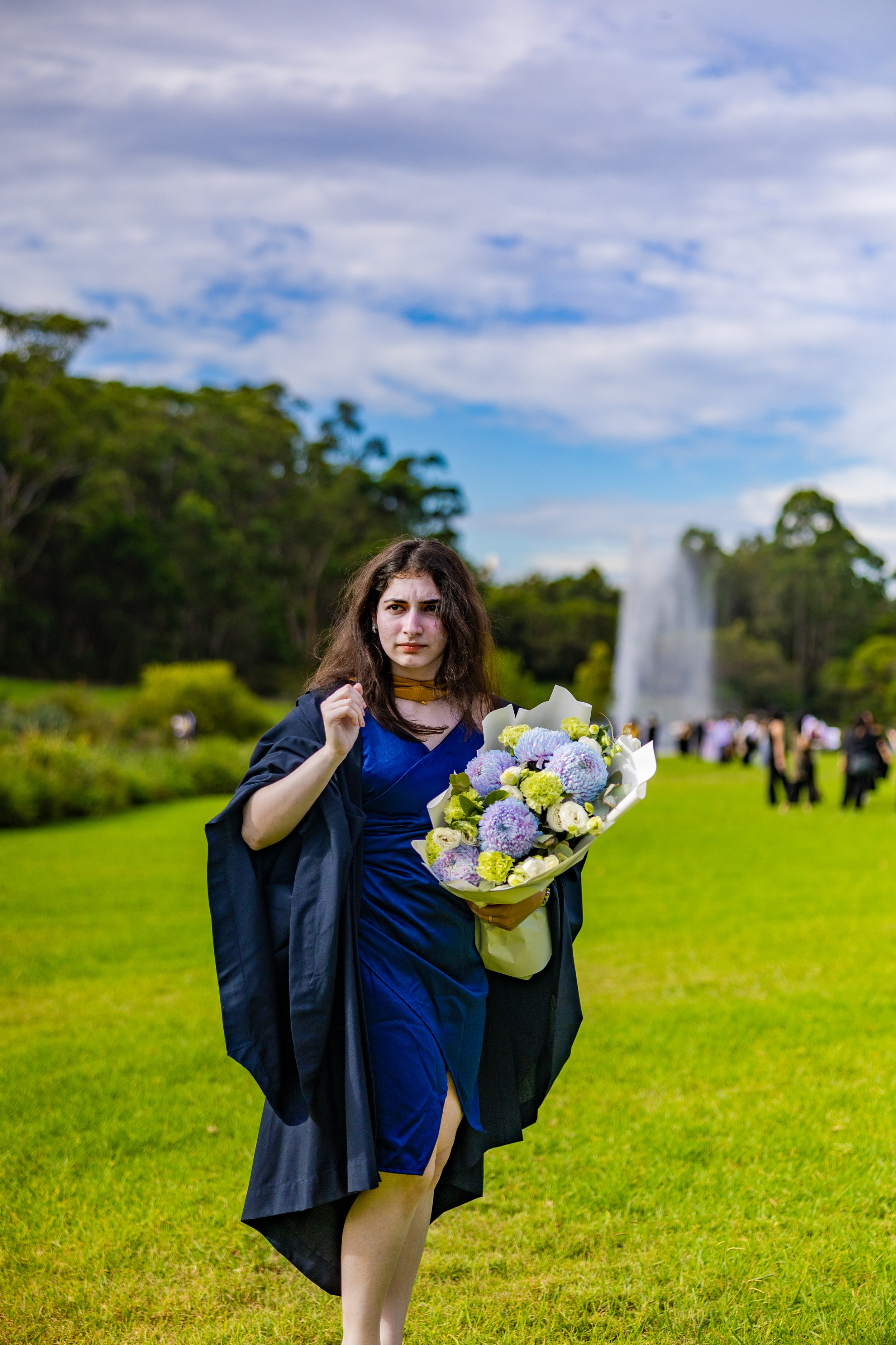 A young woman in a blue dress and black graduation gown holding a bouquet of purple and yellow flowers, standing on a green lawn with a fountain and trees in the background, under a partly cloudy sky.