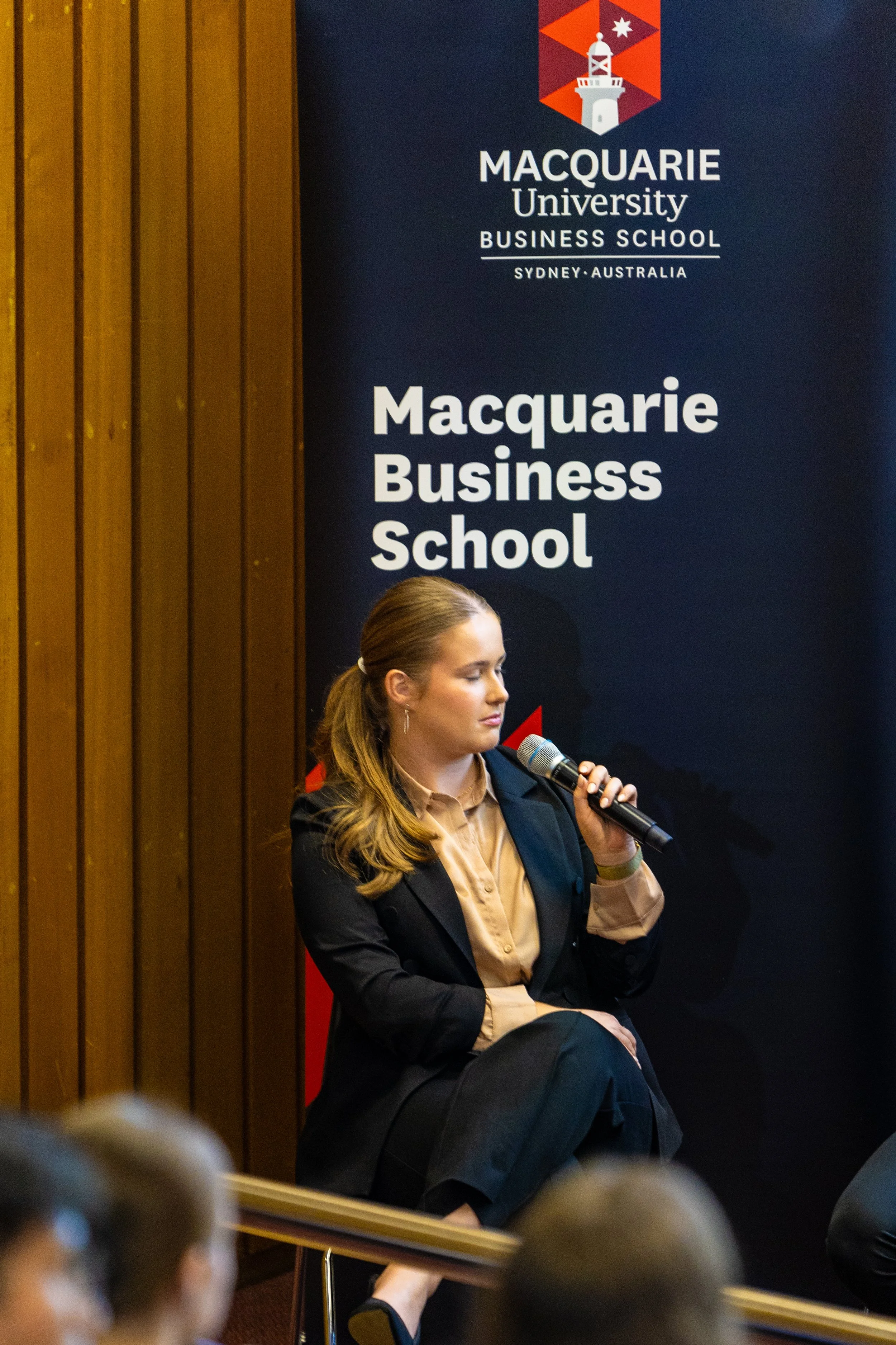A woman in professional attire sitting on a stage at Macquarie University Business School, holding a microphone with her eyes closed, possibly speaking or waiting to speak, with a Macquarie University sign in the background.