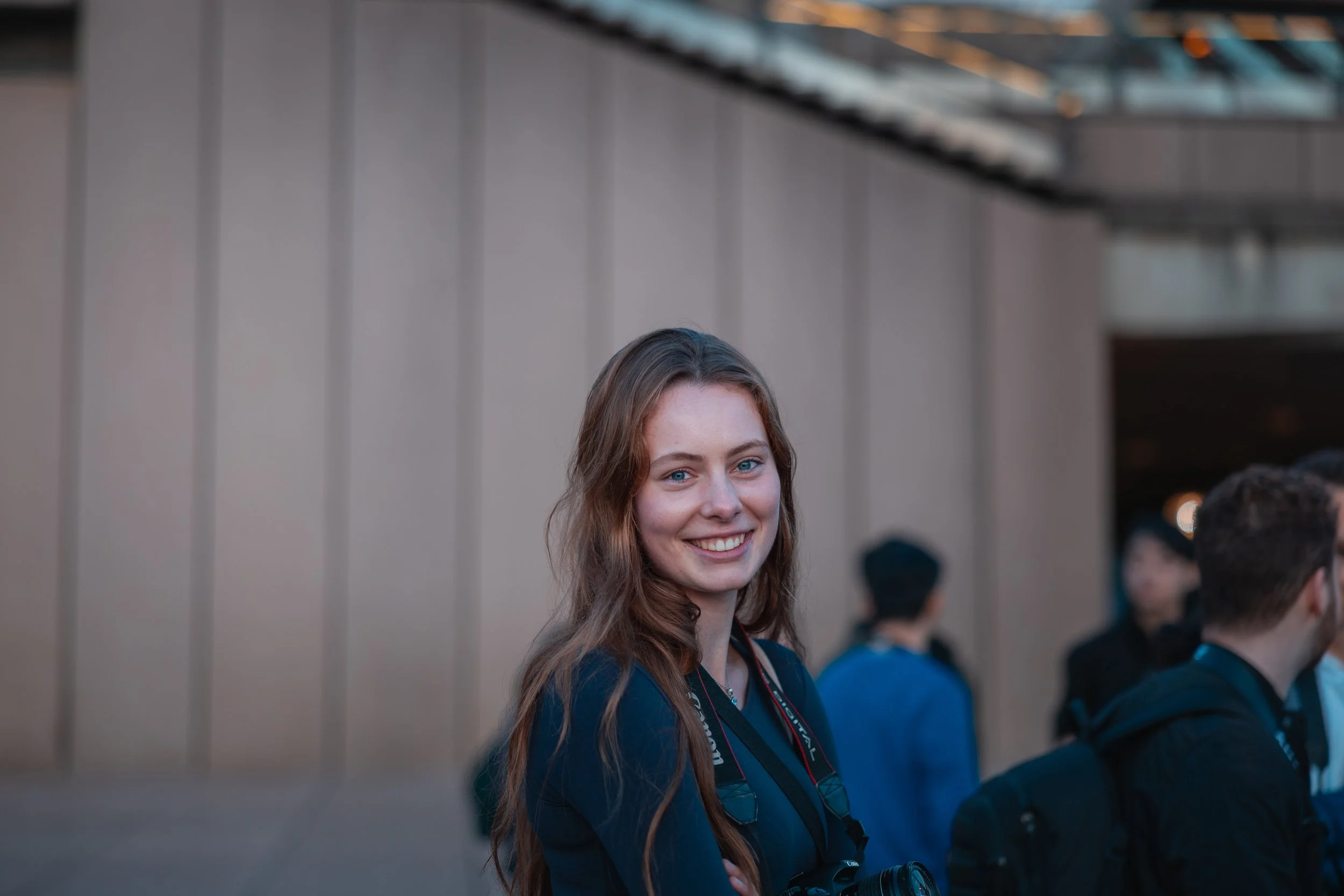 A young woman with long brown hair and blue eyes, smiling, standing outdoors among other people, some with backpacks, in a setting with a beige wall and a glass ceiling.