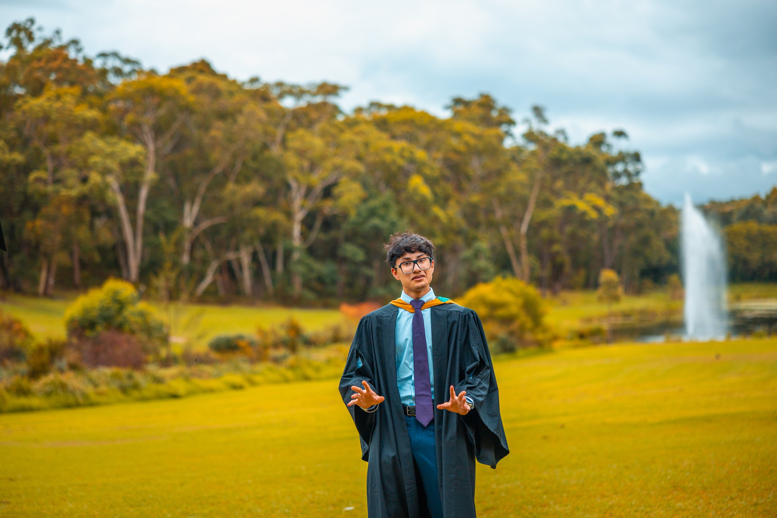 A young man in graduation gown and cap standing in a park with trees and a fountain in the background.