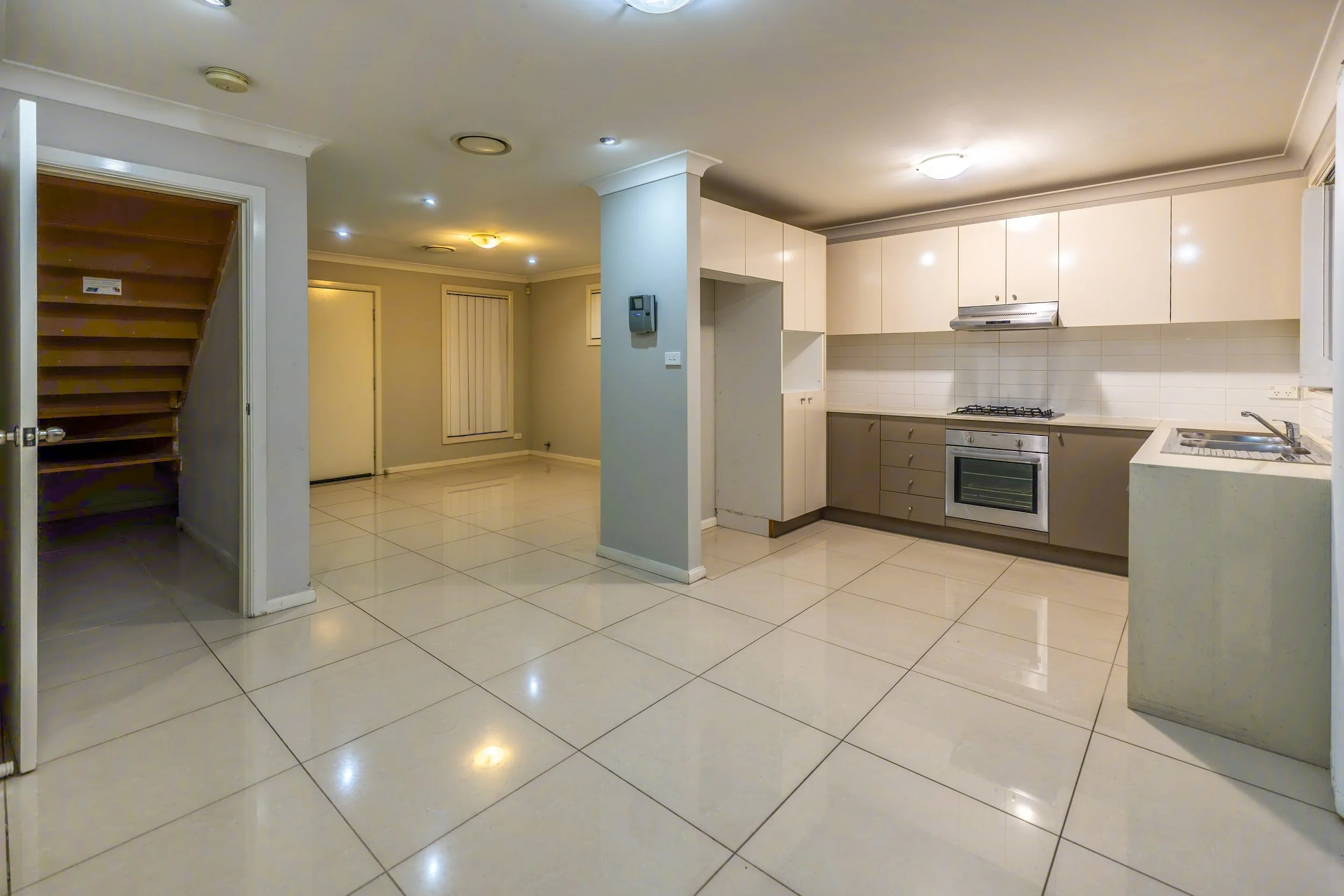 Empty kitchen with white cabinets, gray lower cabinets, tiled backsplash, stainless steel oven, stove, range hood, and a stainless steel sink; adjacent open space with tiled floor and a staircase to the left.