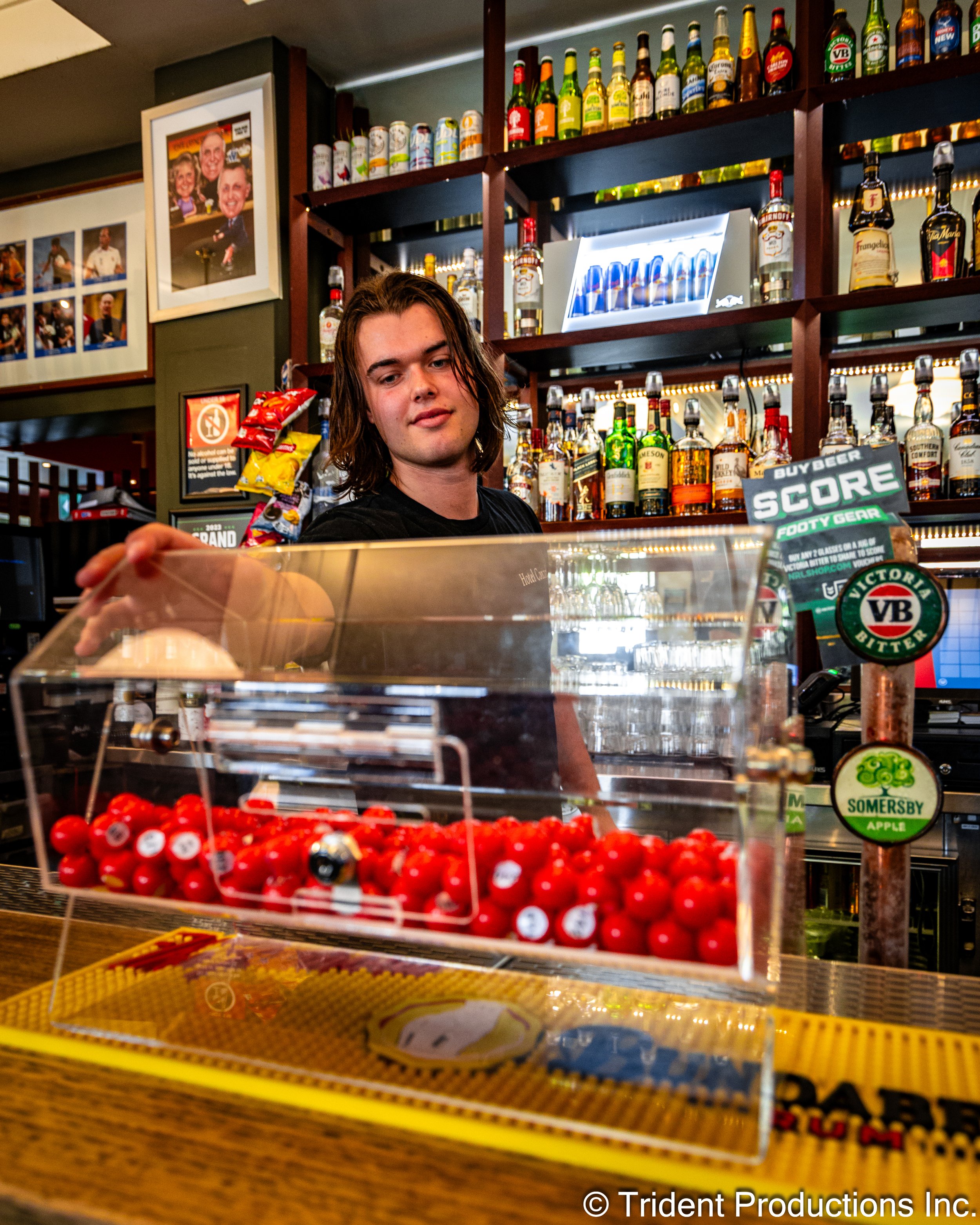 A young man with shoulder-length hair behind a bar counter, reaching into a transparent container of red balls. The background features shelves of liquor bottles and some framed photos on the wall.