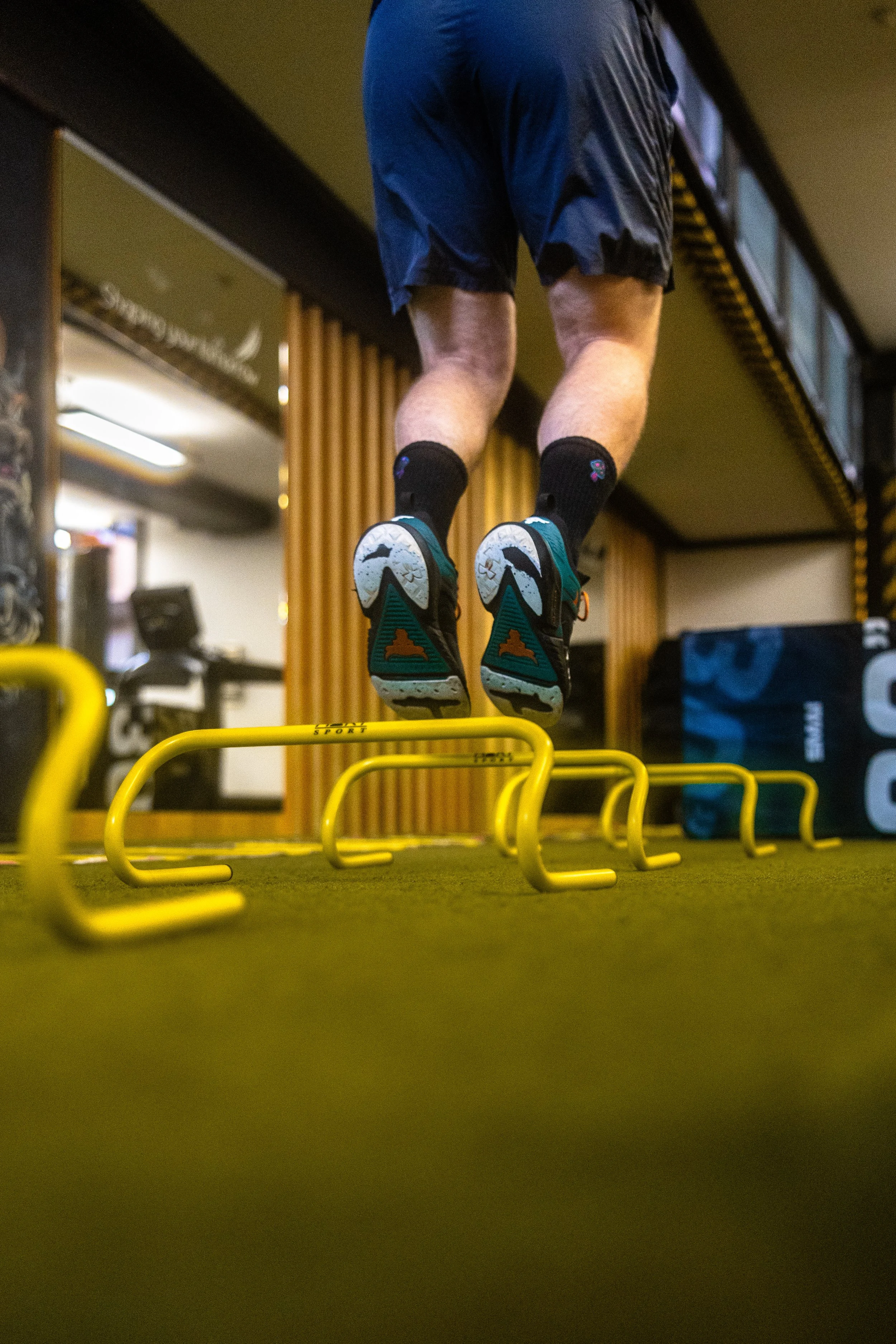 A person jumping over yellow hurdles in a gym or fitness center, wearing athletic shoes, black shorts, and black socks.
