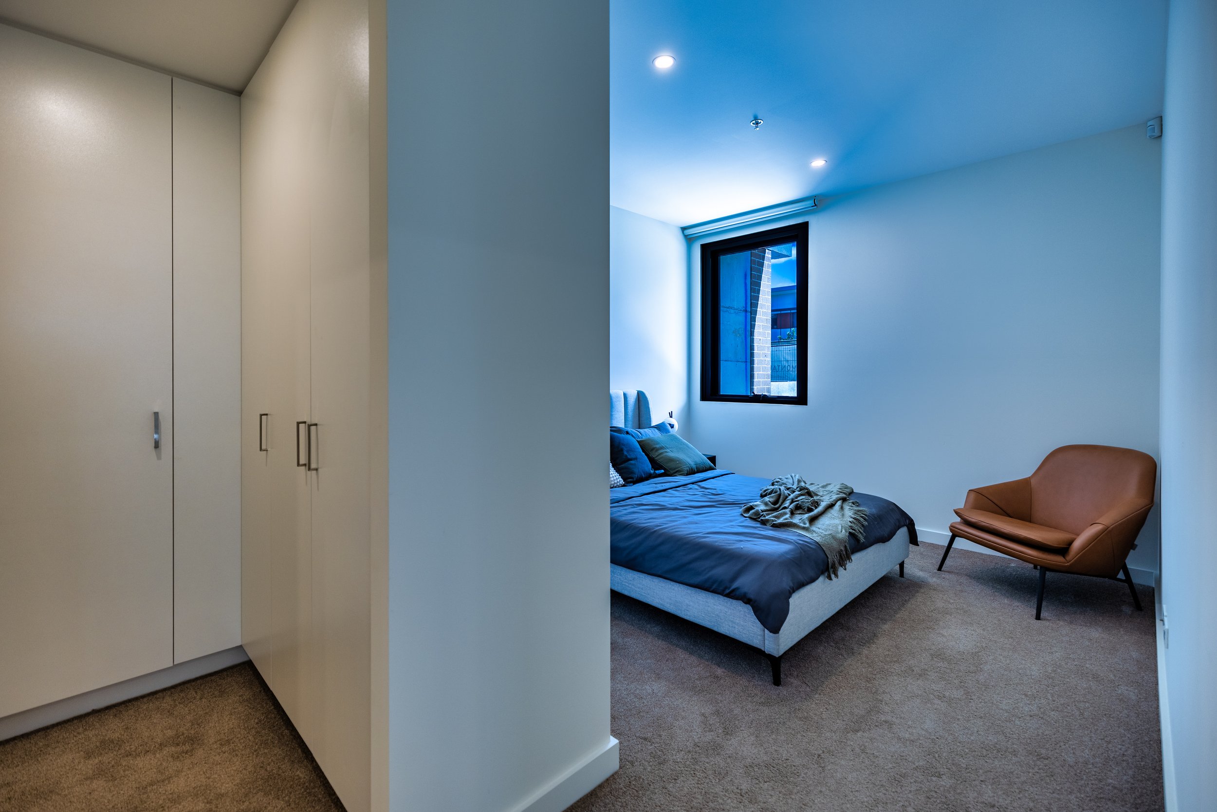 A bedroom with a bed, a window, a brown armchair, closet doors, and ceiling lights.