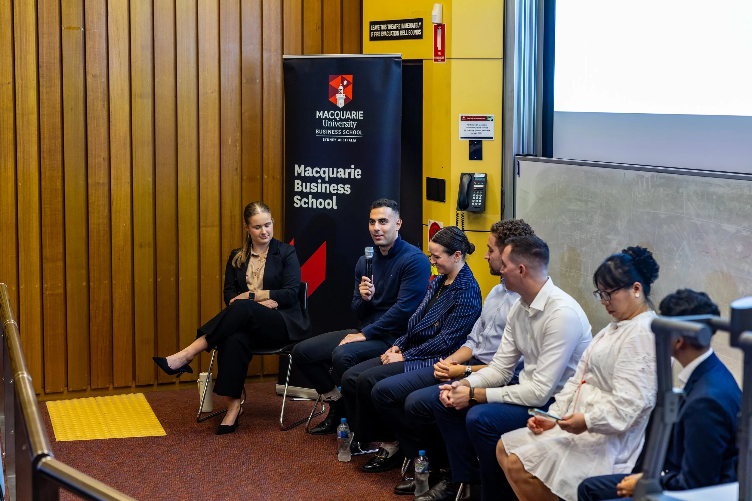 Panel discussion at Macquarie University Business School with six individuals seated in a row, one man speaking into a microphone, with a Macquarie University banner in the background.