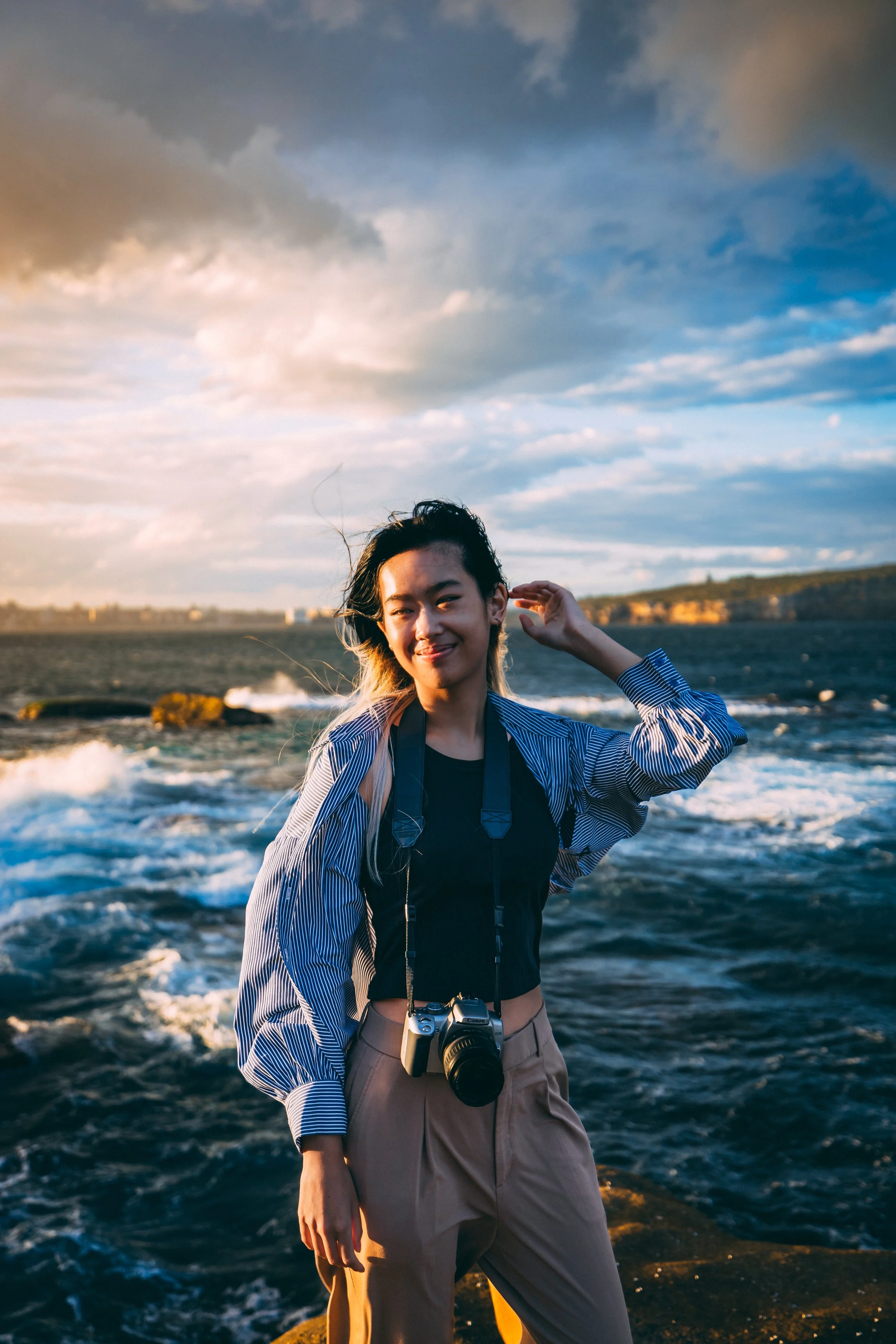 A woman with a camera hanging around her neck standing by the ocean during sunset, smiling with waves and a cloudy sky in the background.