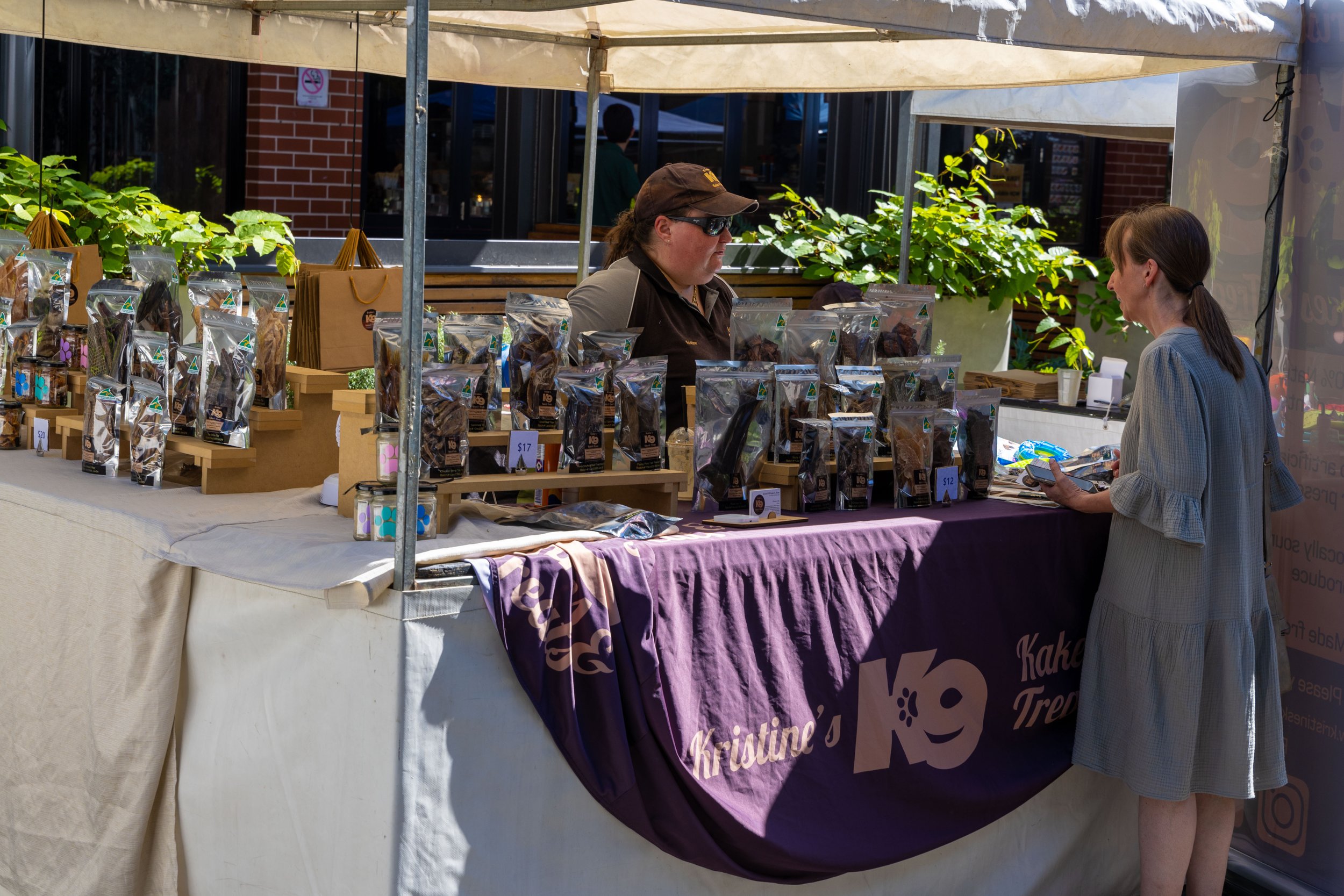 A woman shopping at an outdoor market stall with a purple tablecloth and a purple banner that reads 'Kristine's Kake' or similar text. The stall has packaged baked goods on display and a vendor woman behind the table wearing sunglasses and a cap.