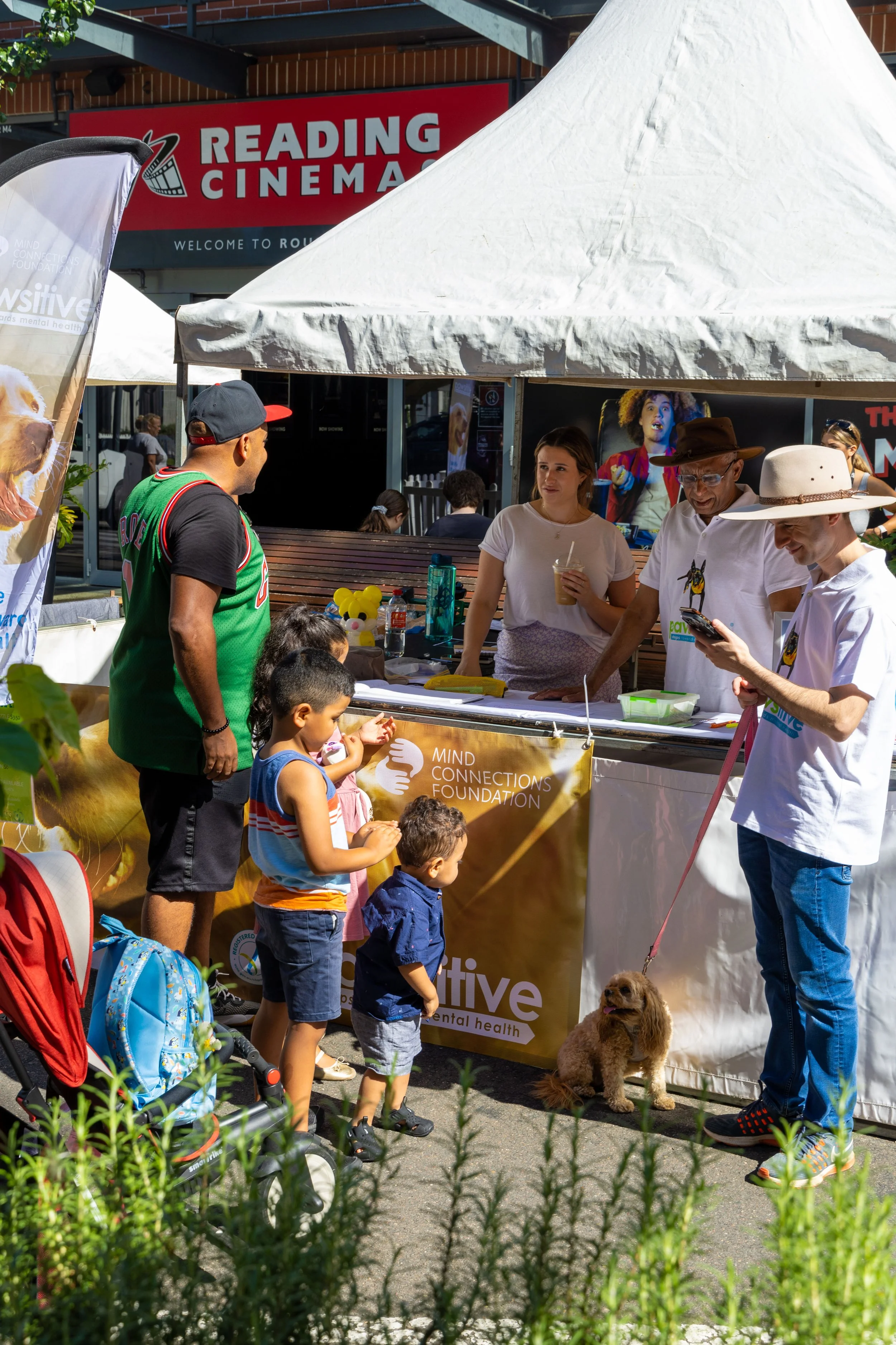 People at an outdoor booth for the Mind Connections Foundation, with children and a dog in front, and a Reading Cinema sign in the background.