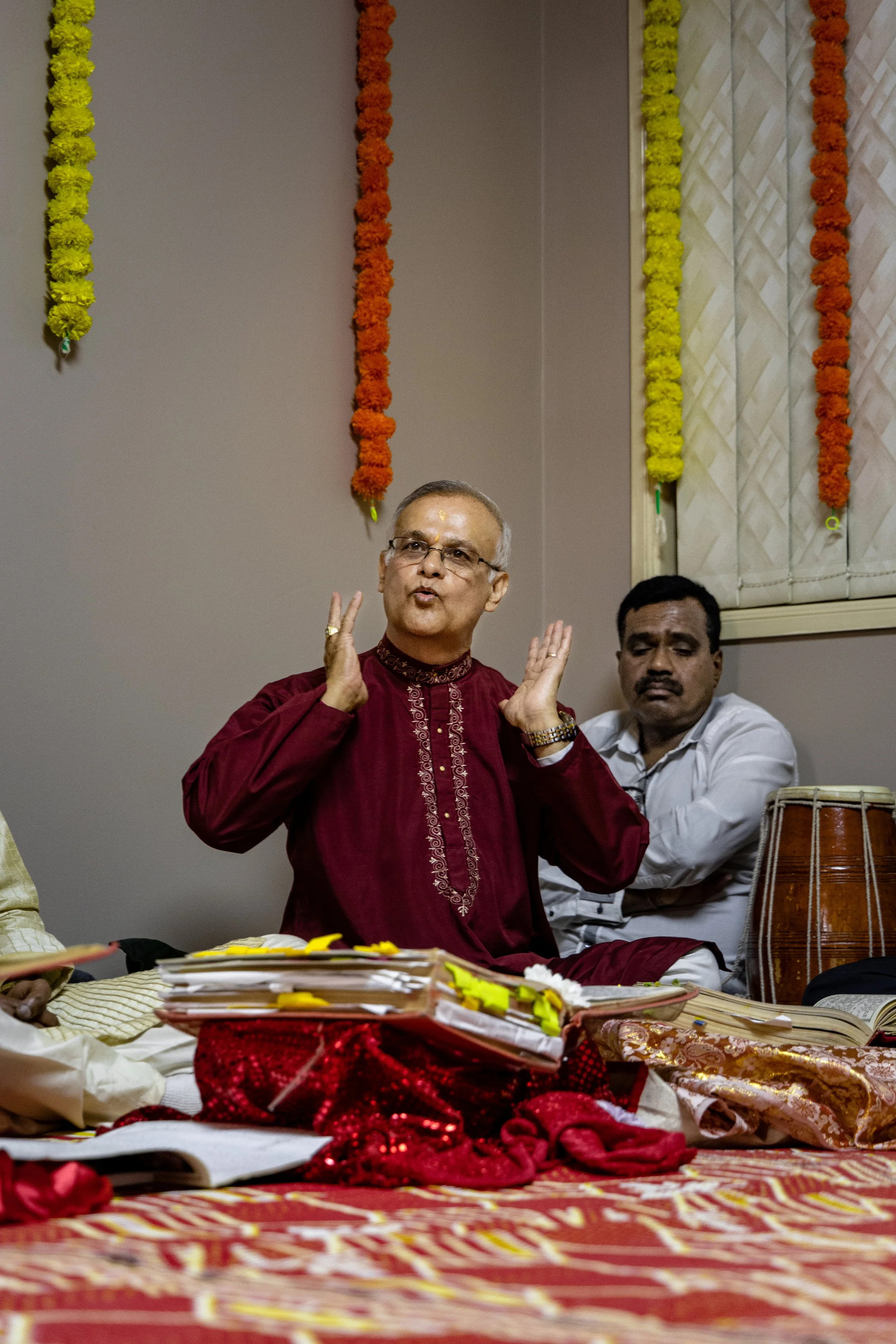 An older man with glasses and yellow markings on his forehead is speaking or singing, wearing a maroon traditional Indian kurta. He is sitting on a decorated floor with a pile of papers and cloths. Behind him, another man in a white shirt is seated, 