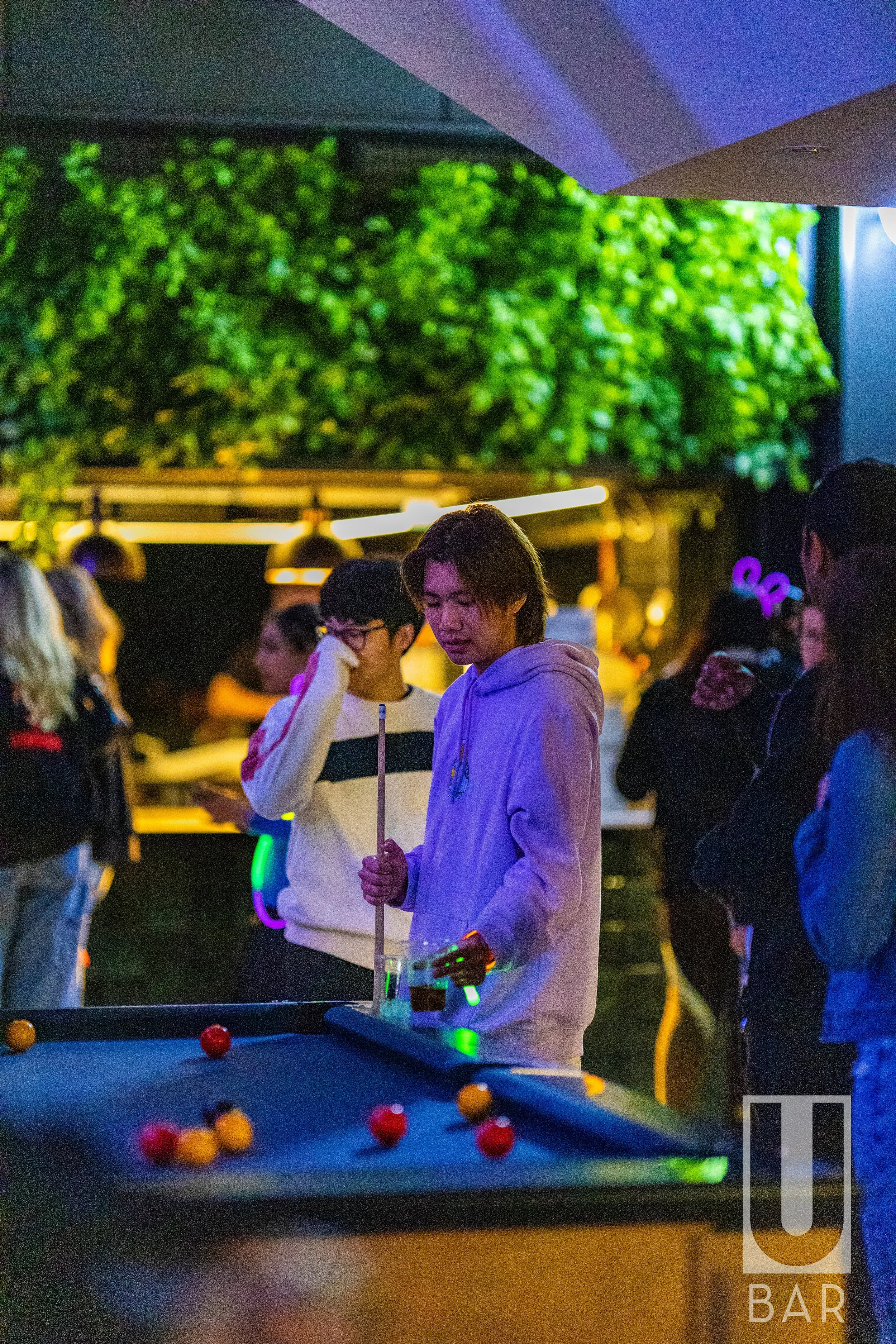 A group of young people at a bar or nightclub, with one person playing pool under colorful lighting, and others gathered around in the background.