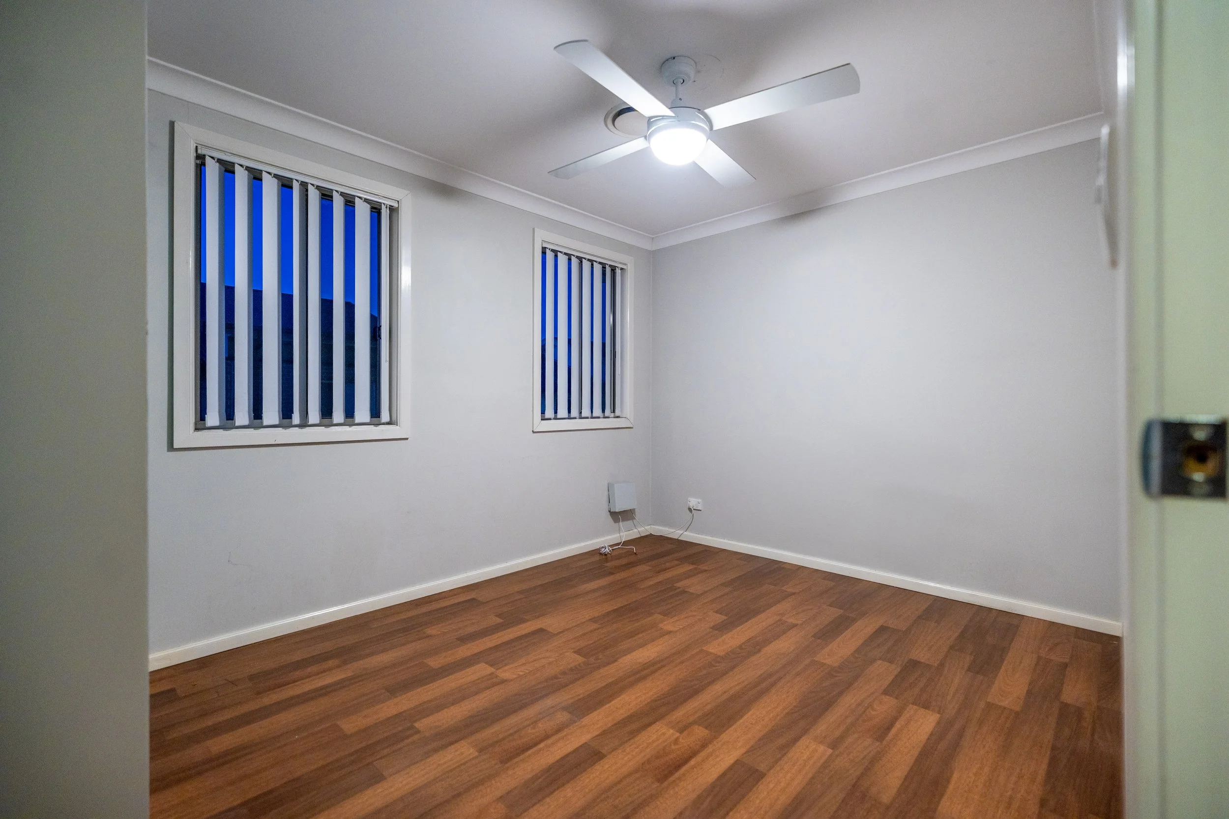 Empty room with white walls, two windows with vertical blinds, wooden floor, ceiling fan with light, and wall outlet.