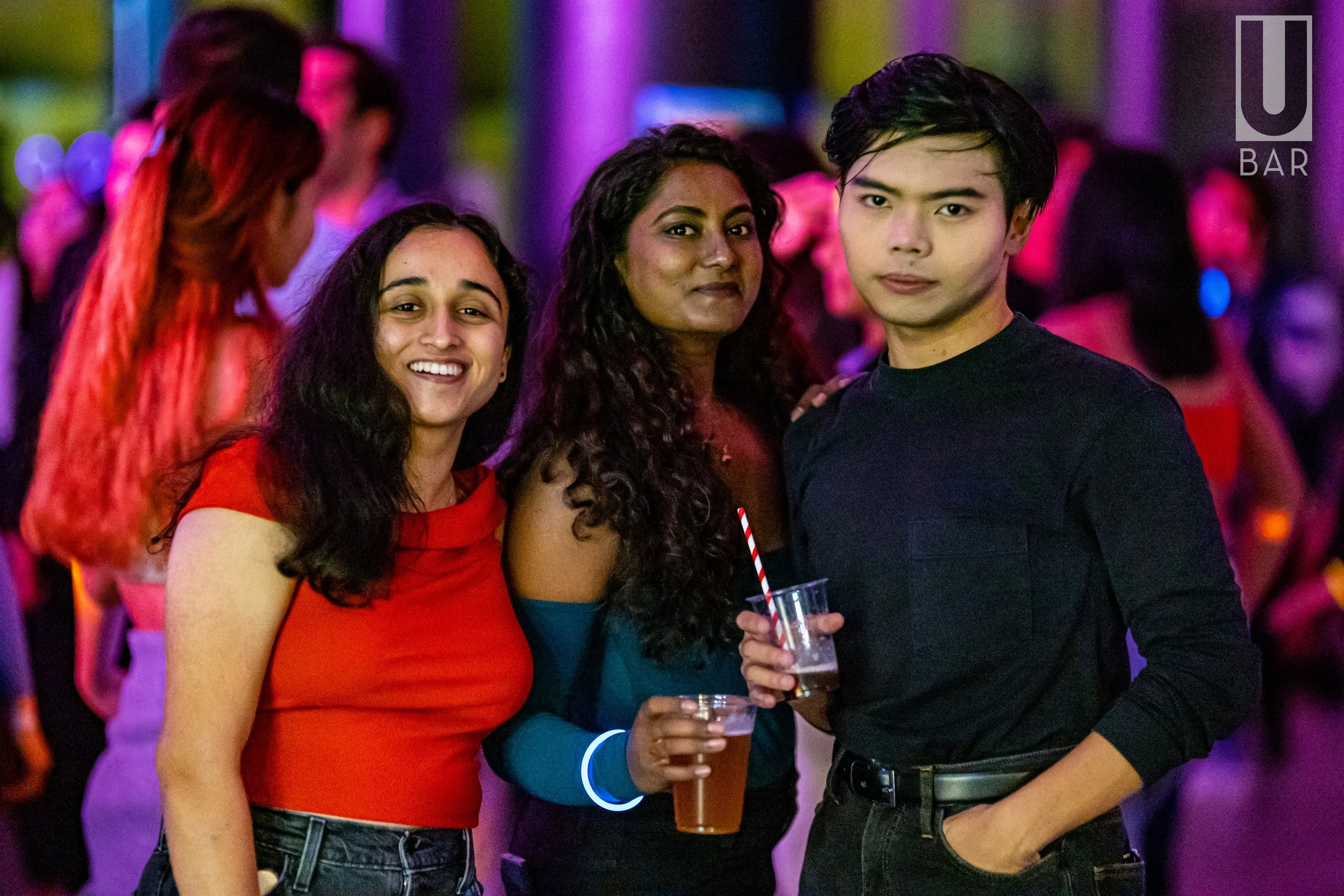 Three friends at a lively bar, holding drinks, smiling, with colorful lights and other patrons in the background.