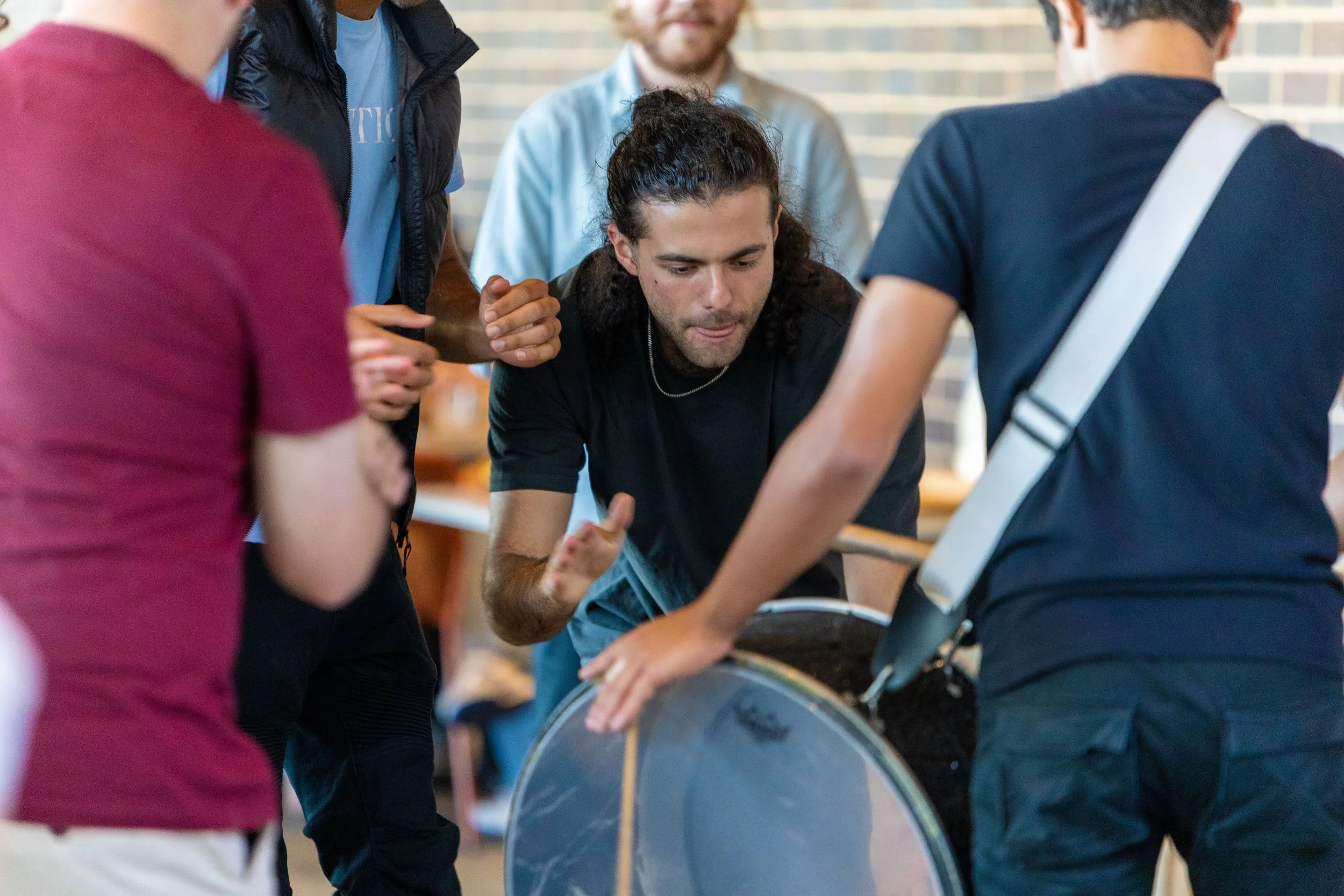 Group of people gathered around a large drum, with one person leaning over it and others supporting or observing, in an indoor setting.