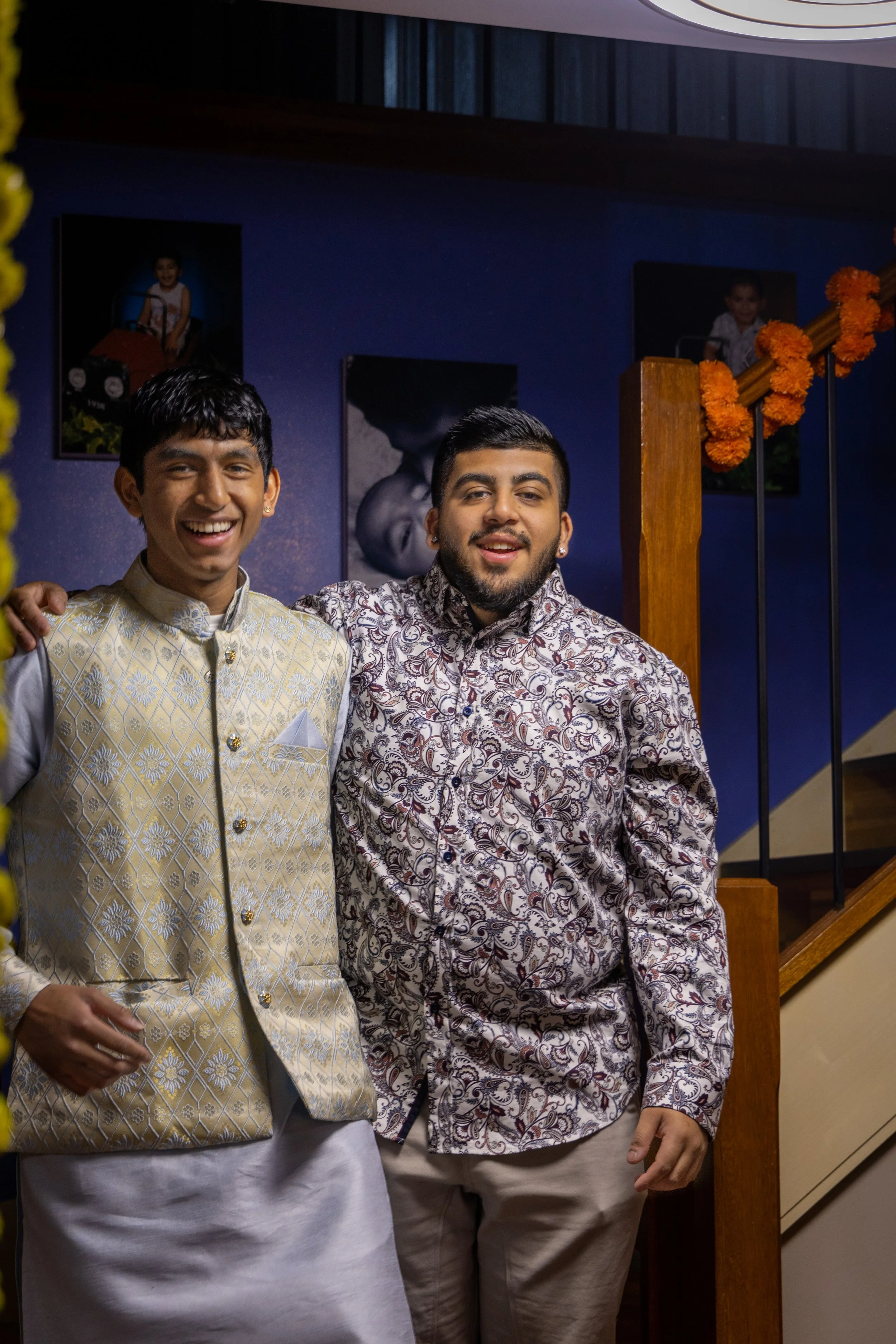 Two young men standing together indoors, smiling at the camera, with a blue wall, framed photos, and orange marigold decorations in the background.