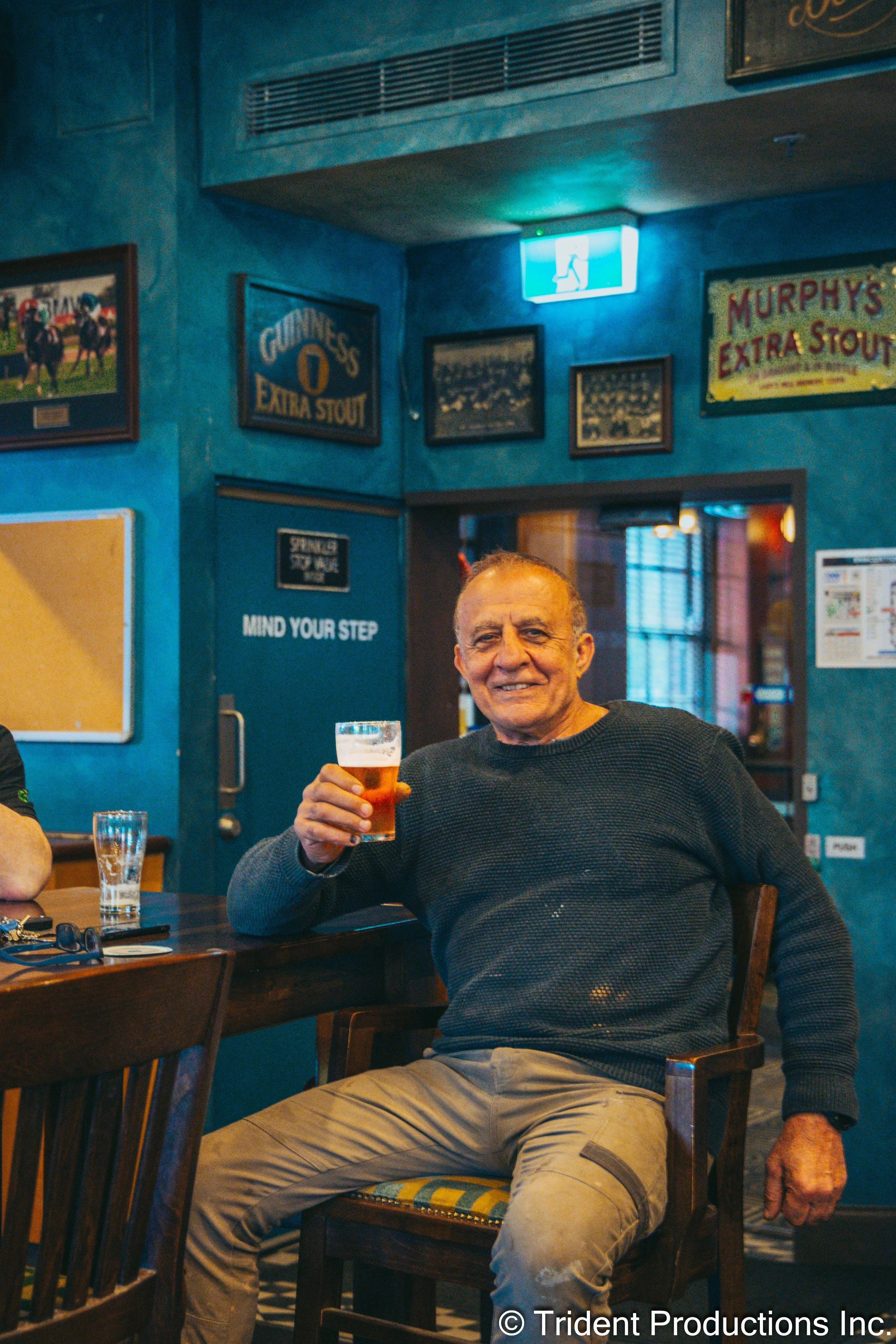 An older man in a black sweater sitting at a bar, smiling, holding a pint of beer in a pub with blue walls decorated with framed pictures, signs, and an emergency exit sign overhead.