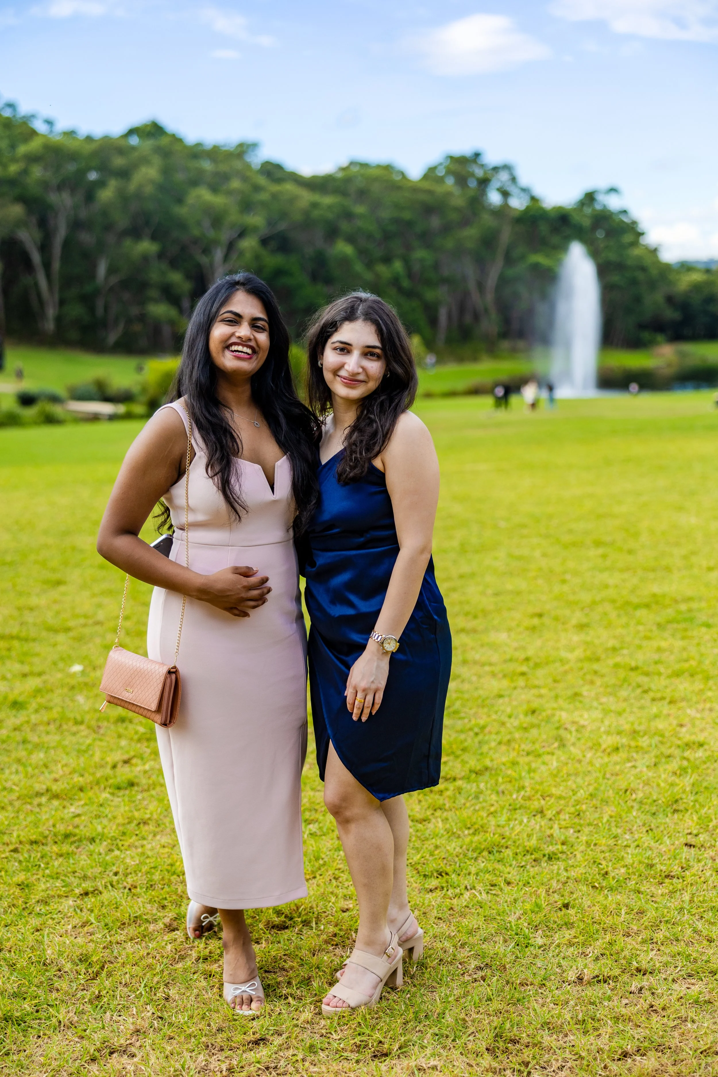 Two women standing on grass in a park with a fountain and trees in the background, smiling at the camera.