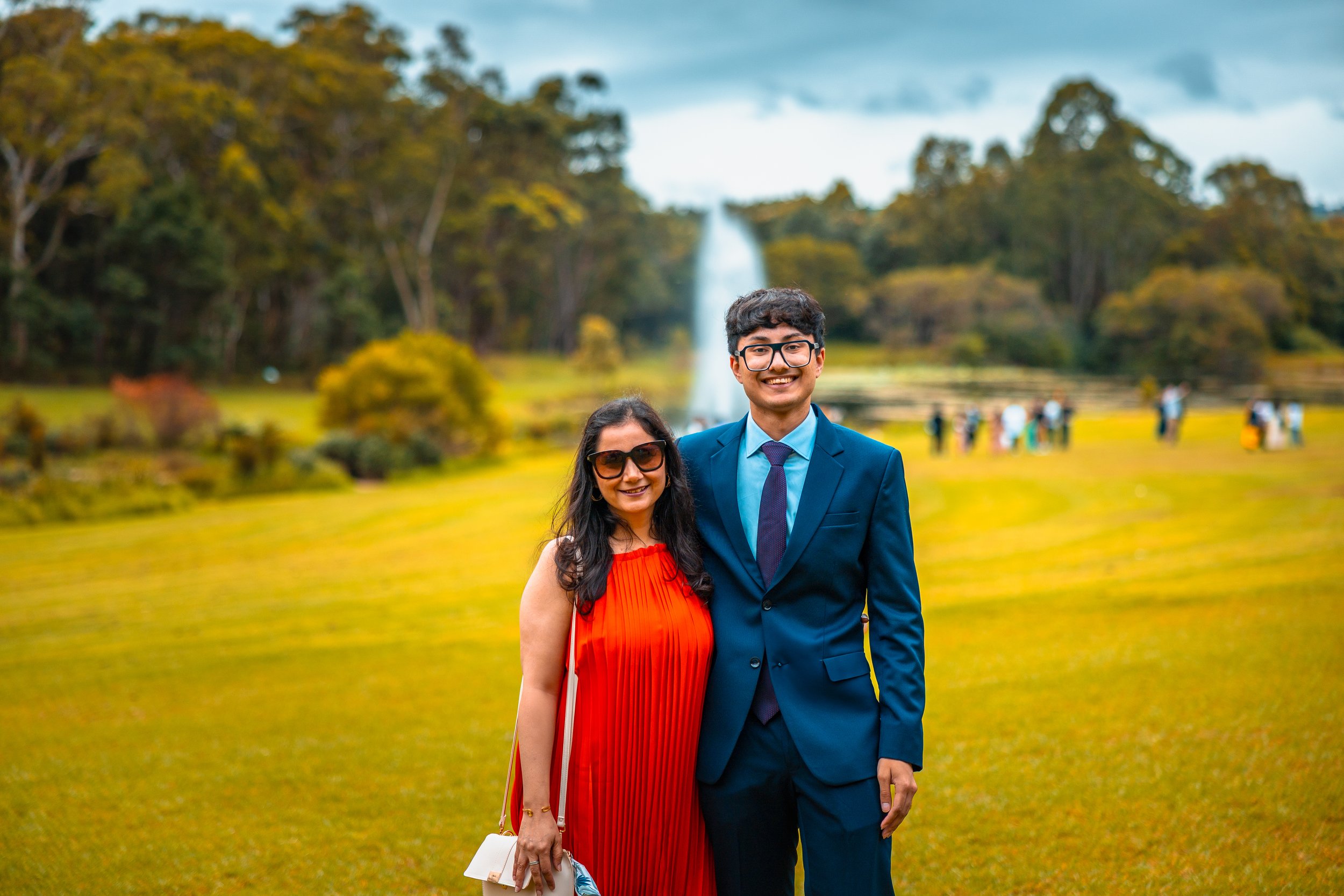 A woman and a man standing together outdoors in a park, smiling at the camera. The woman is wearing a red dress and sunglasses, and the man is wearing a blue suit with a tie. There are trees and a fountain in the background.
