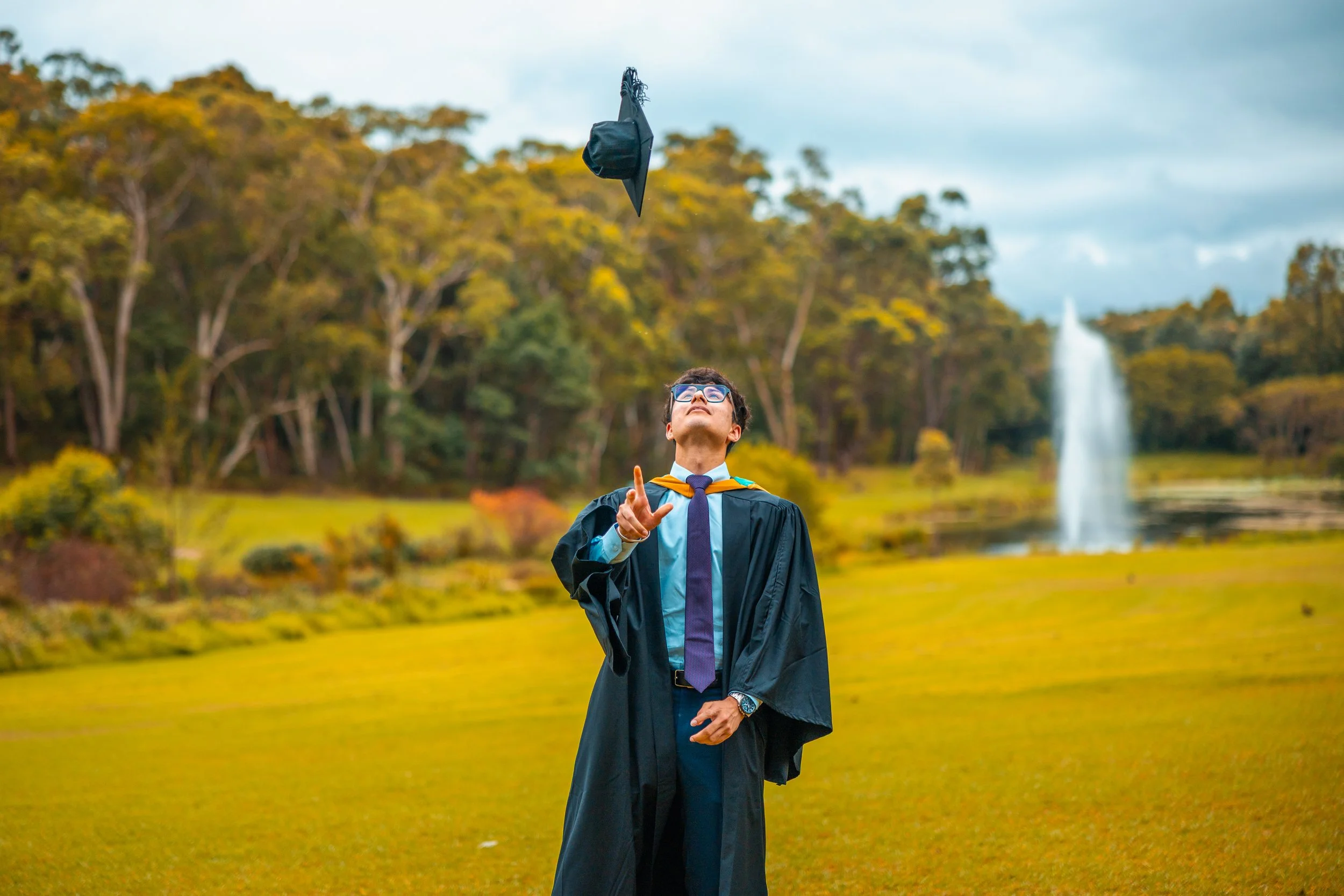 A man in graduation attire standing on a grassy area, tossing his cap into the air, with trees and a water fountain in the background.