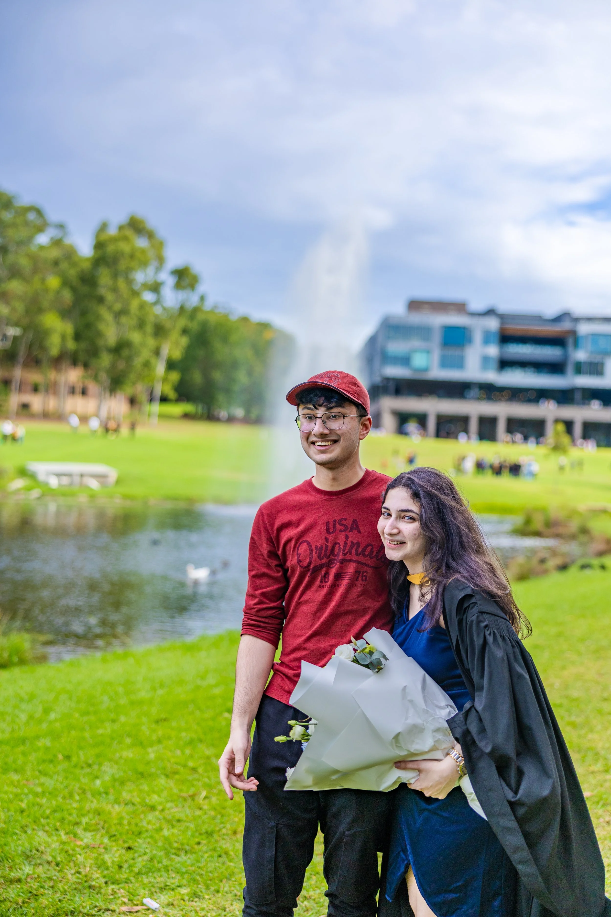 Two young adults, a man and a woman, smiling outdoors near a pond with a fountain, trees, and a modern building in the background. The woman is holding a bouquet of flowers and is dressed in a graduation gown.