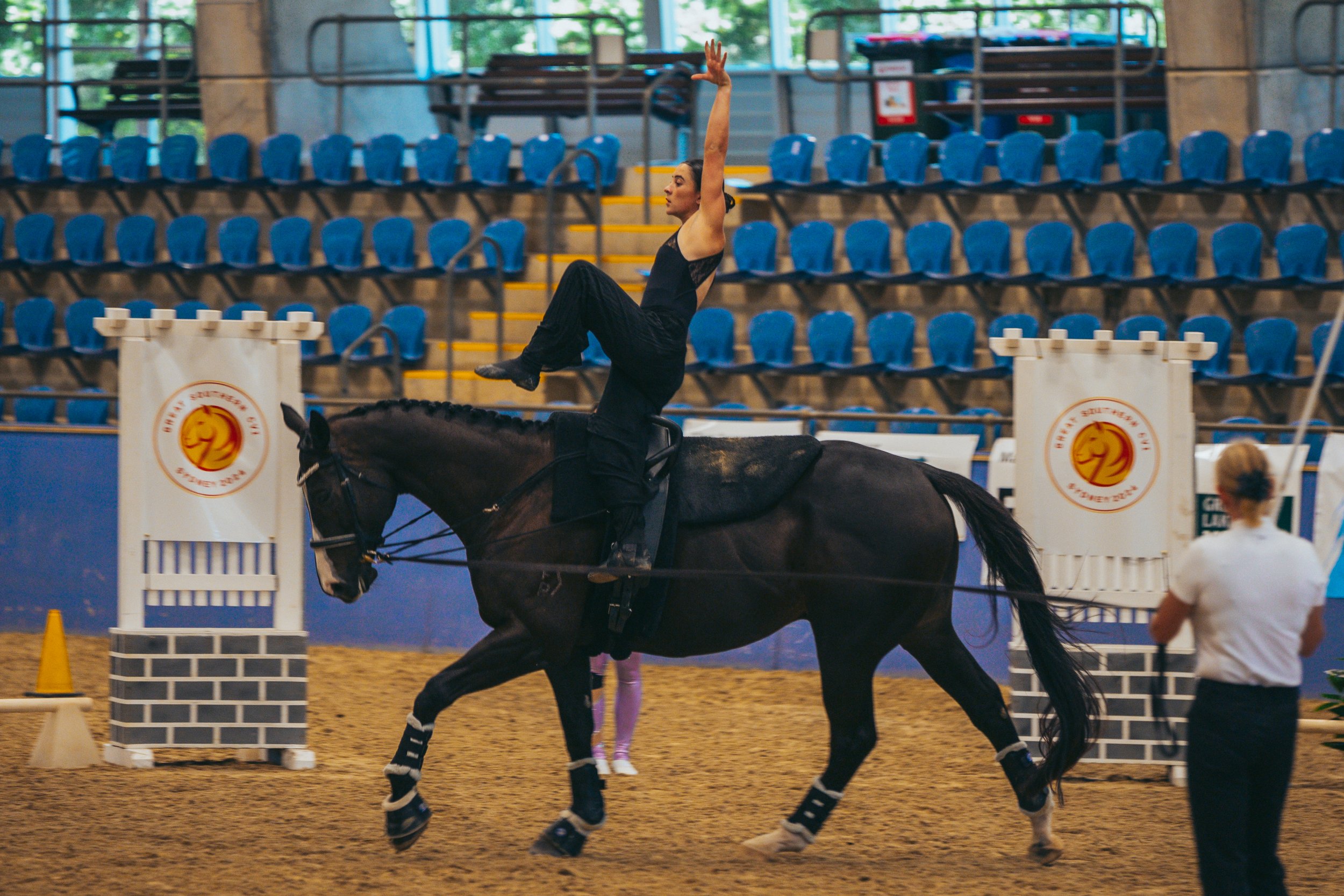 A female equestrian performer standing on a black horse in an indoor arena, with her right arm raised and left arm holding onto the horse, surrounded by blue seats and judges or officials watching.