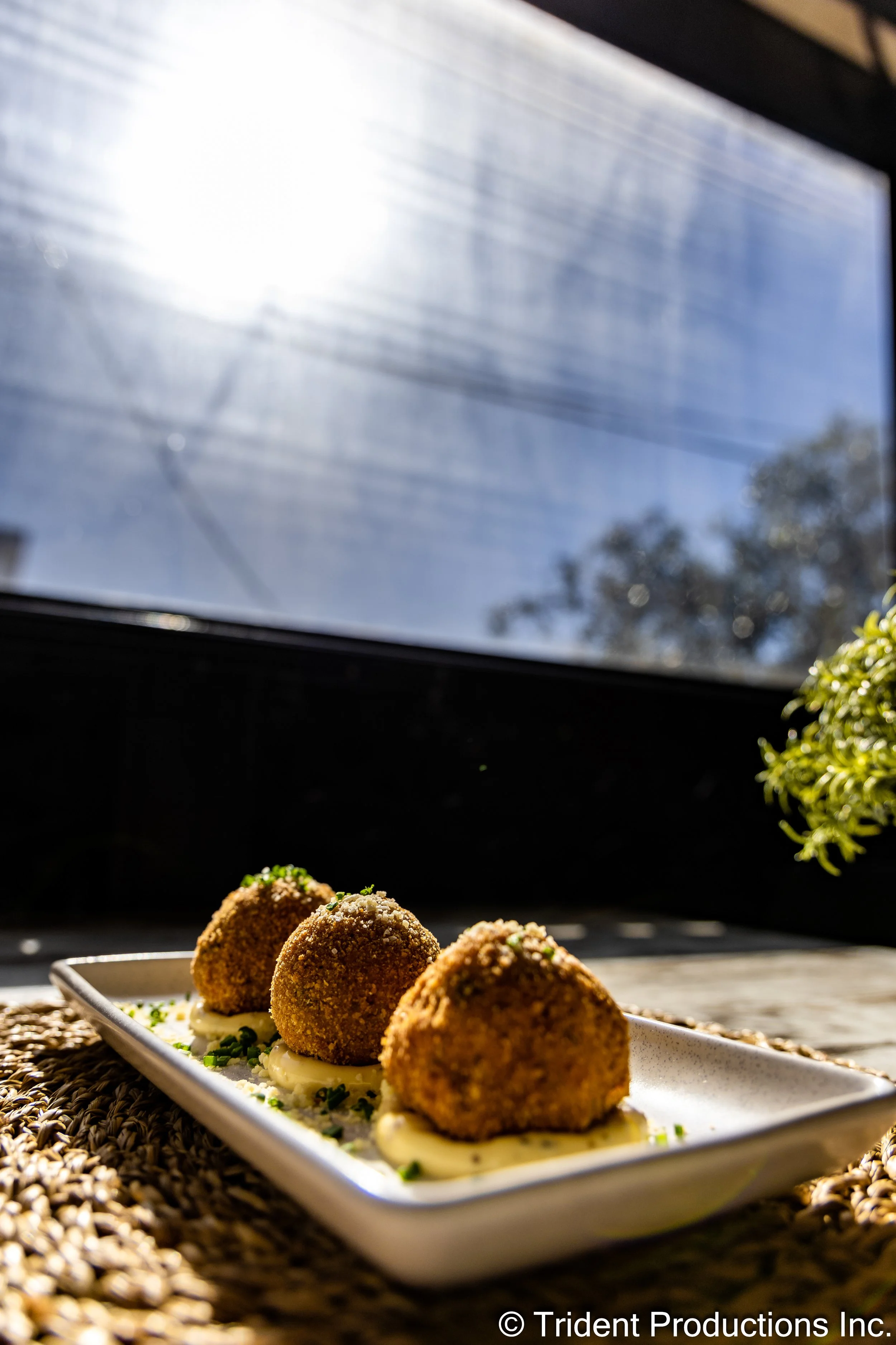 Three golden-brown breaded croquettes served on a rectangular white plate, garnished with chopped herbs, placed on a woven mat near a window with sunlight and a blue sky in the background.