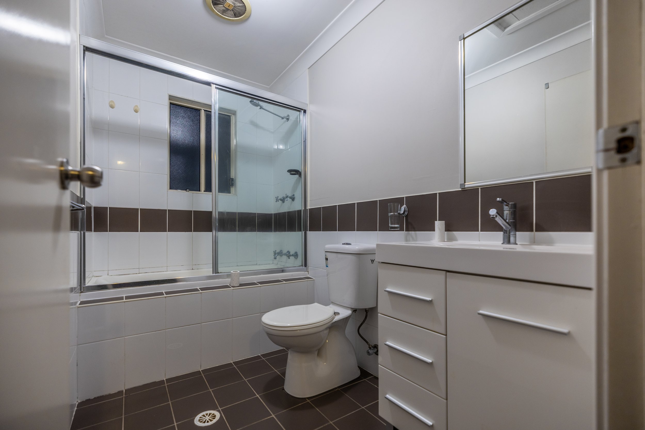 A small bathroom with a bathtub and a sliding glass door shower, a toilet, a white cabinet with a sink, a large mirror, and brown tiles on the lower half of the wall.
