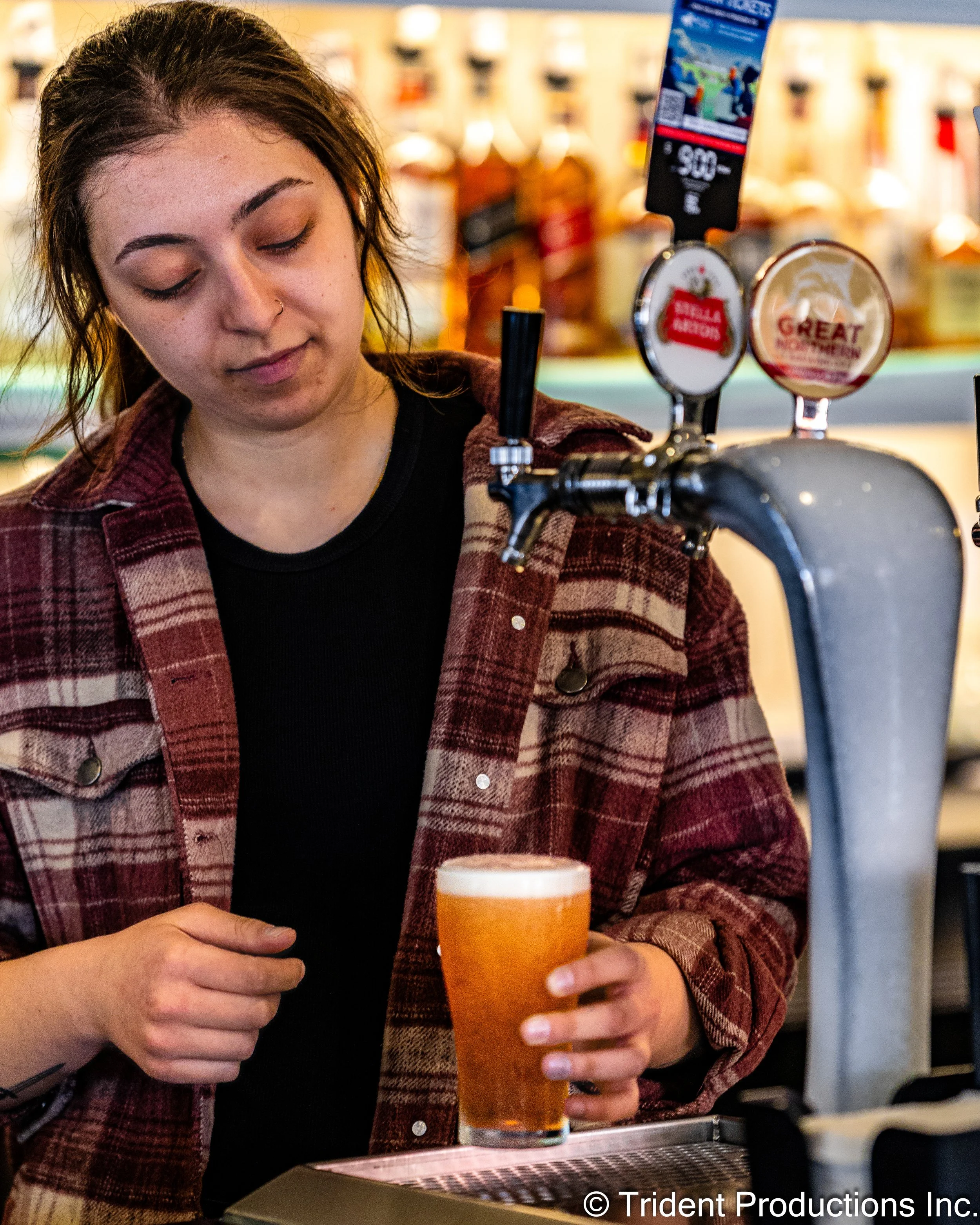 A young woman with dark hair and a nose piercing pours beer from a tap at a bar.