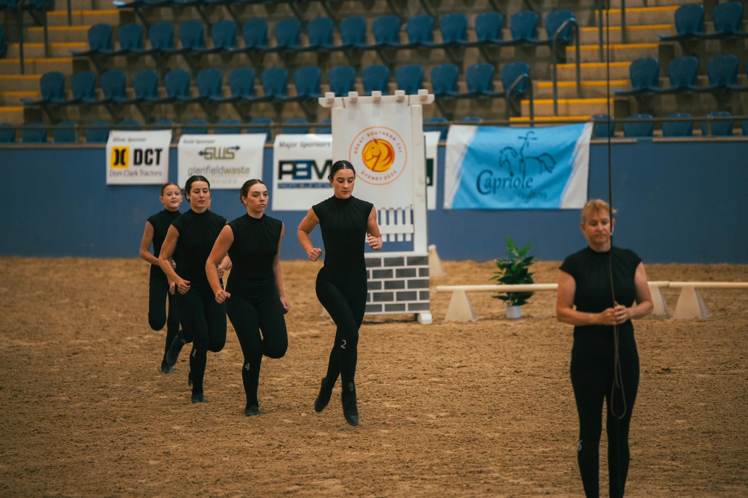 Four women running in an indoor arena with a sandy floor, with one woman holding a leash and standing still, behind them are blue empty seats and advertising banners.