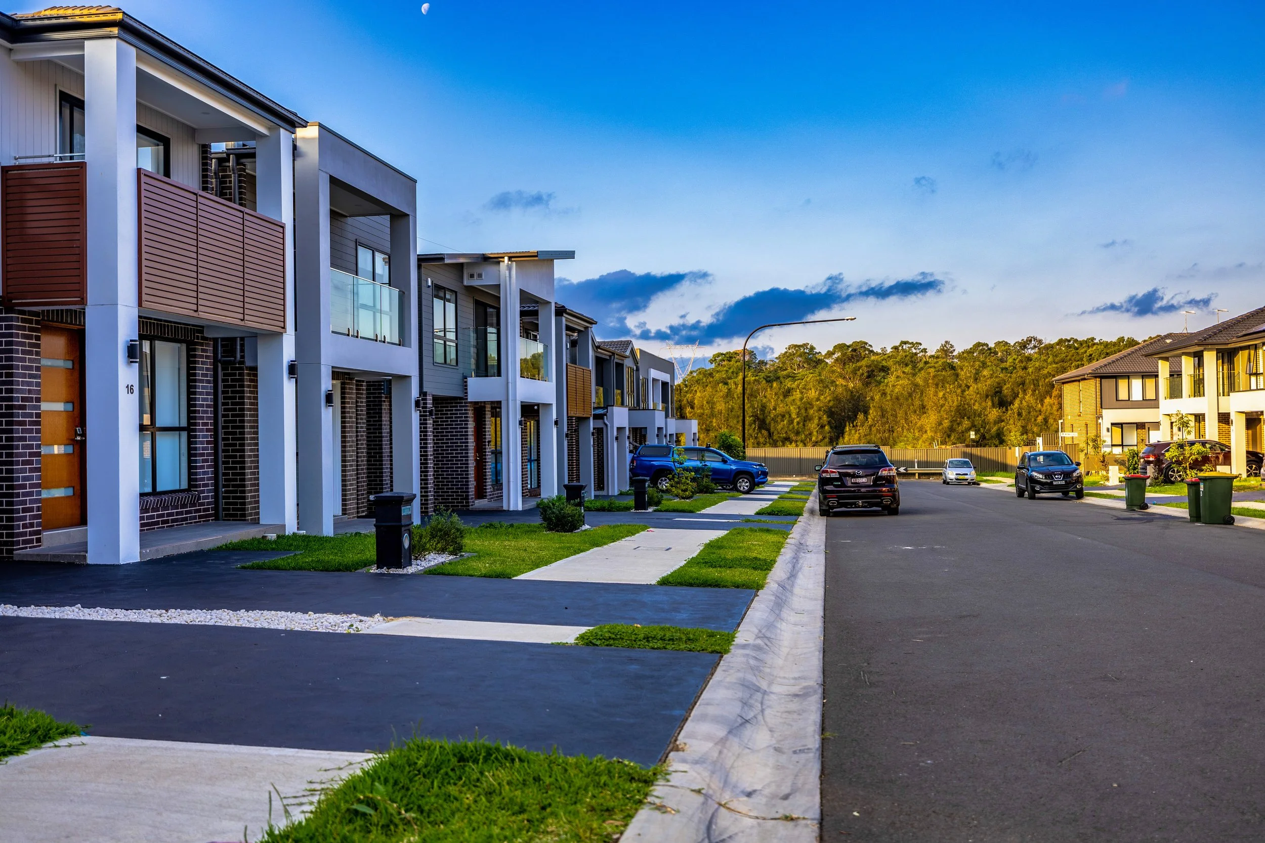 Modern residential street with row of townhouses, parked cars, green lawns, and a clear sky at sunset.