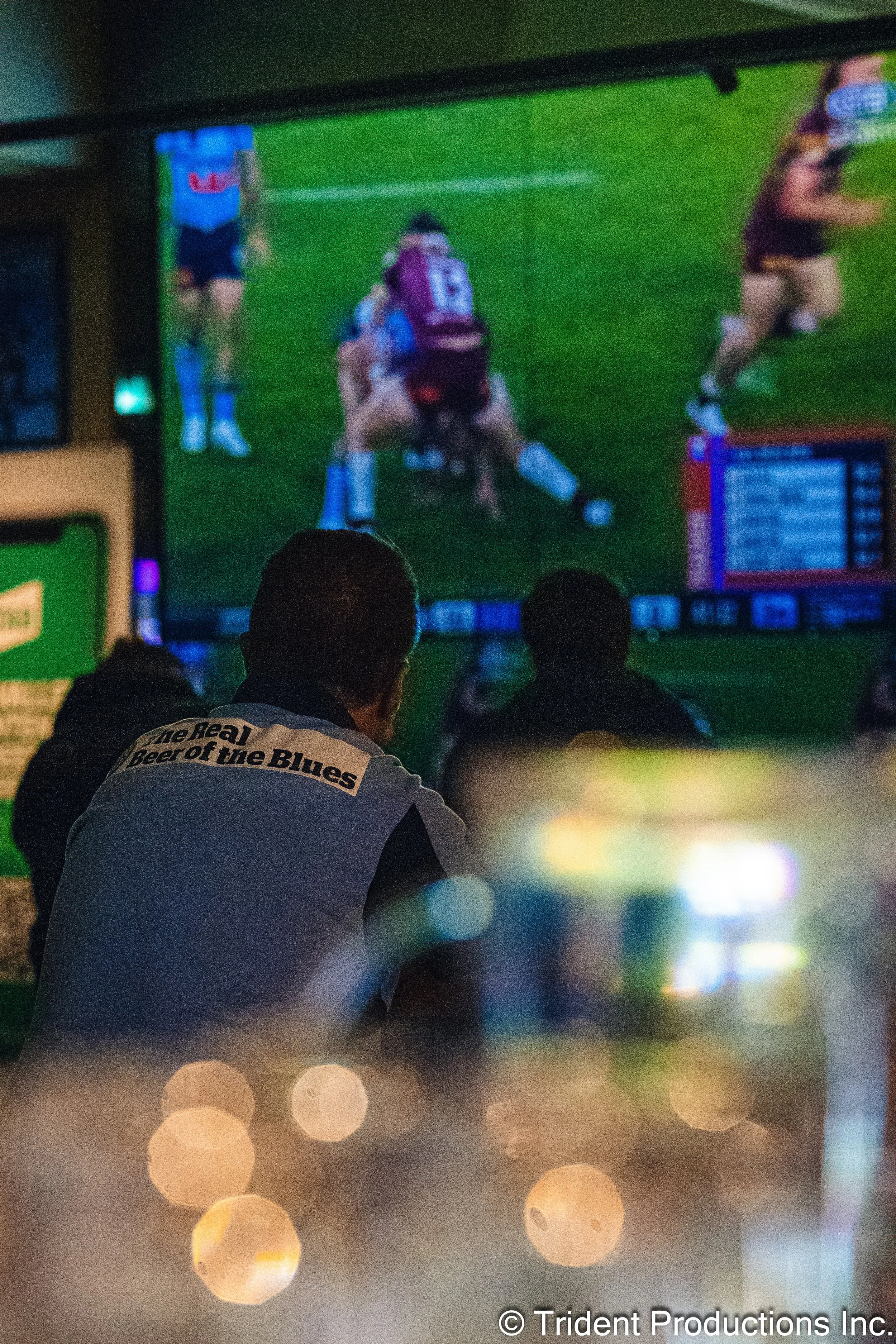 People watching a televised rugby game in a dimly lit bar, with a man wearing a shirt that says "The real Beer of the Blues" in the foreground and a large screen displaying players on a grassy field.