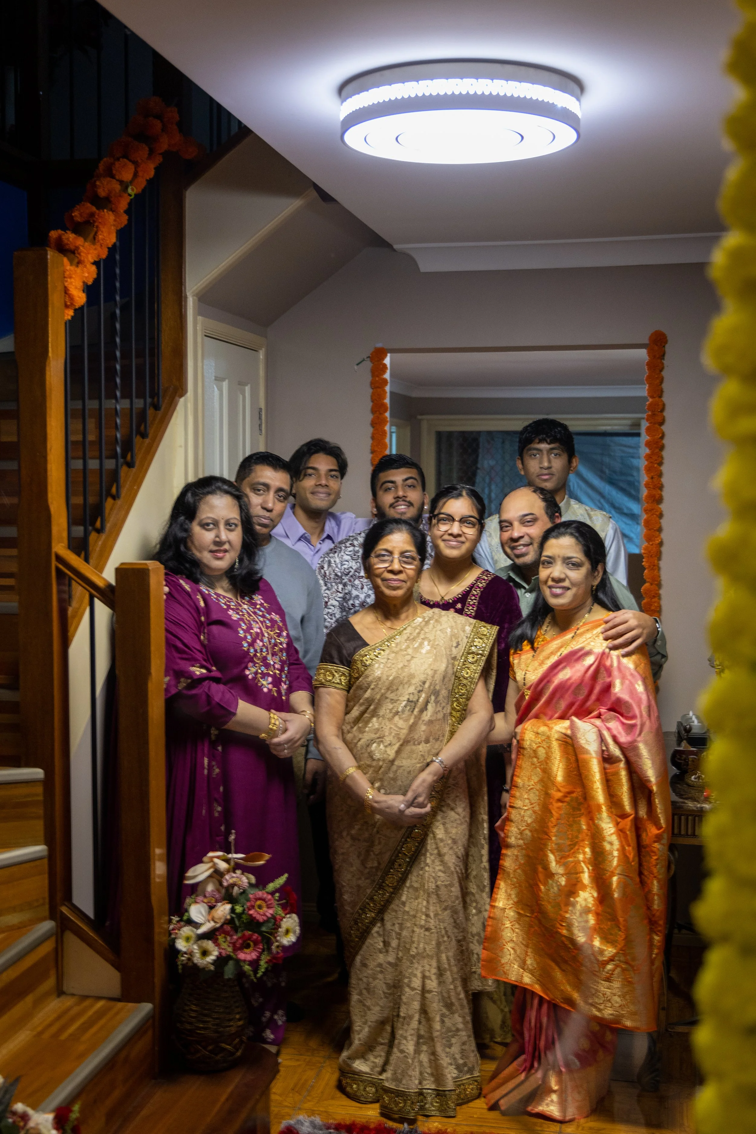A family of nine people dressed in traditional Indian attire posing together inside a home decorated with orange marigold garlands for a celebration.