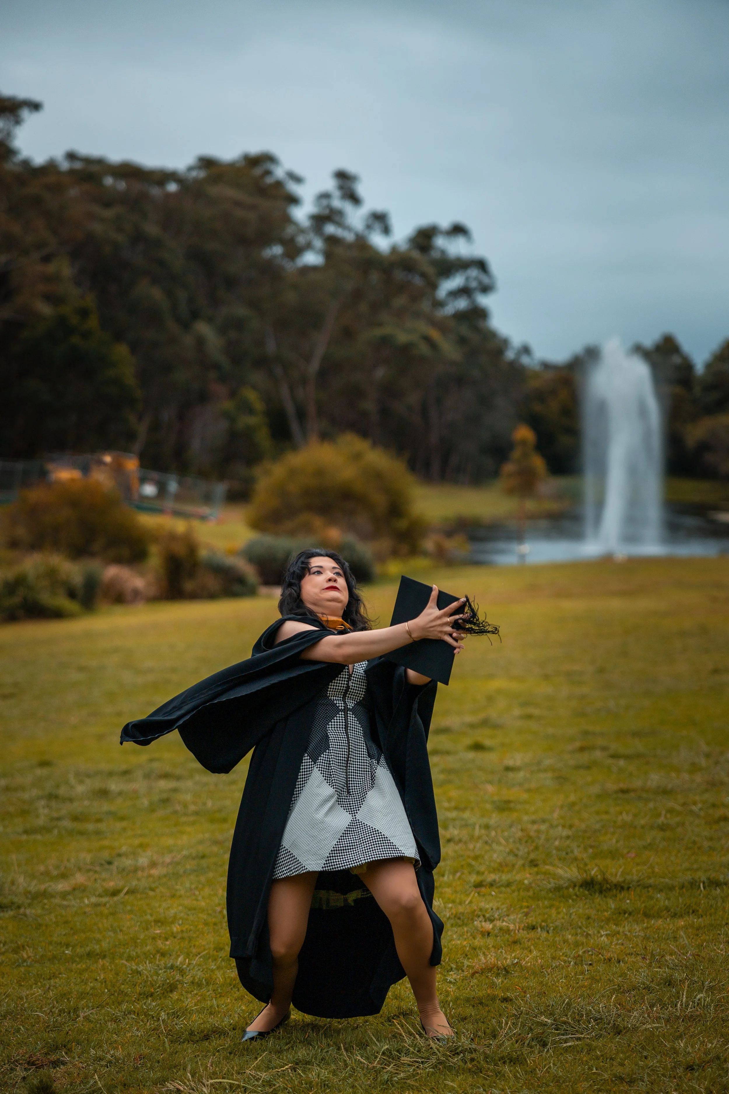 Woman in academic regalia taking a selfie outdoors with a waterfall and trees in the background.