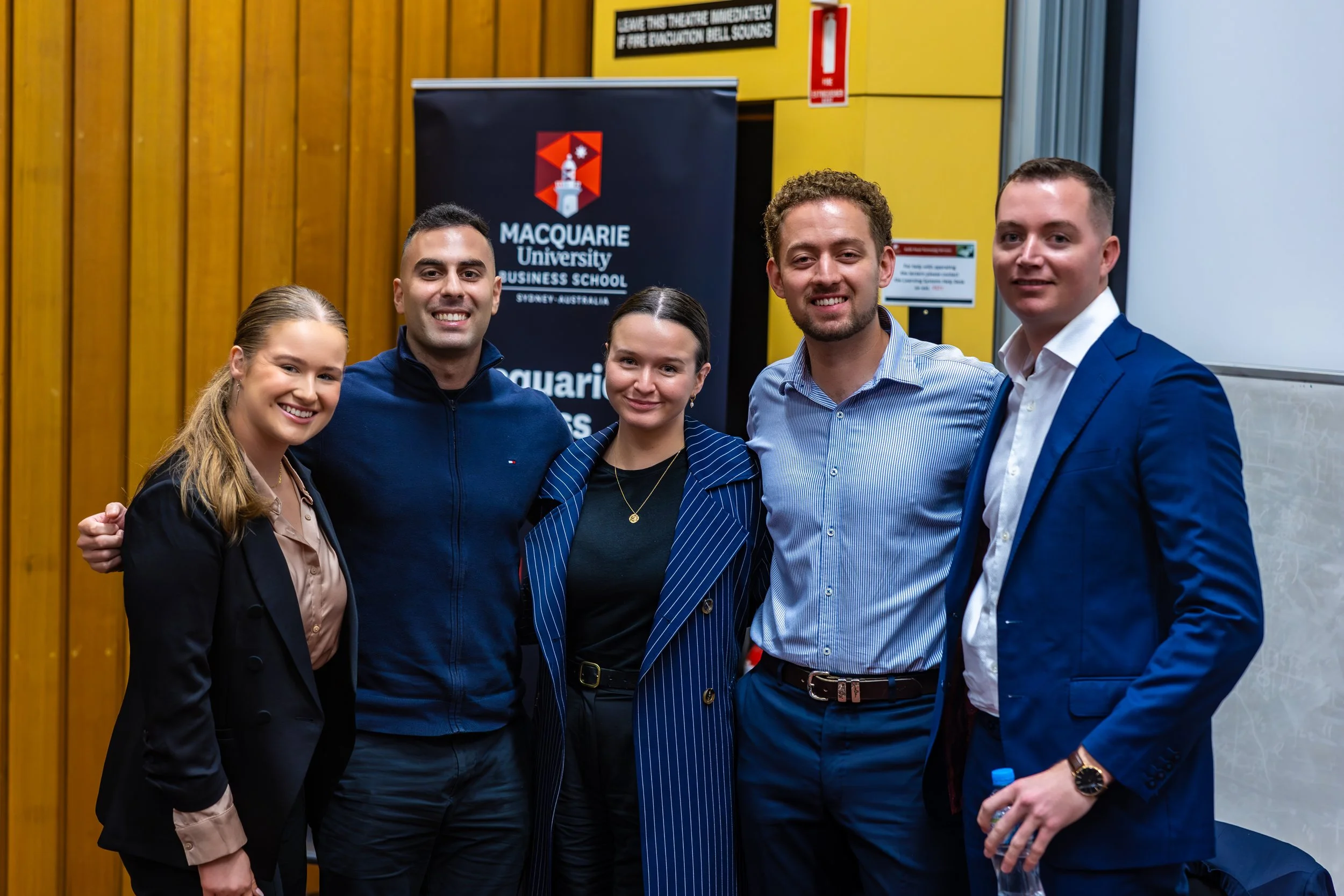 Group of five young professionals smiling for a photo at an event, standing in front of a Macquarie University Business School banner.
