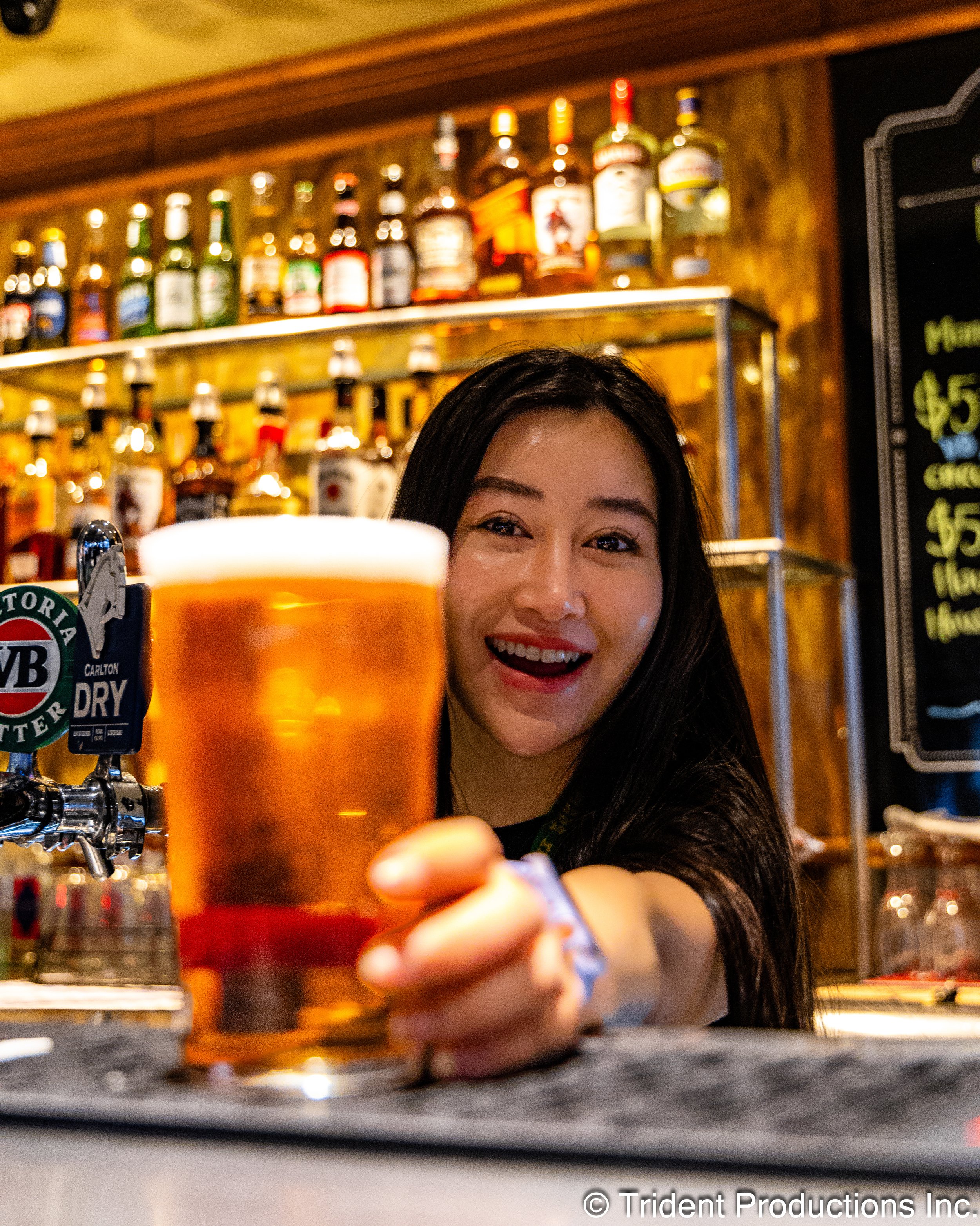 A woman with dark hair smiling and holding a glass of beer at a bar, with bottles of alcohol displayed on shelves behind her.