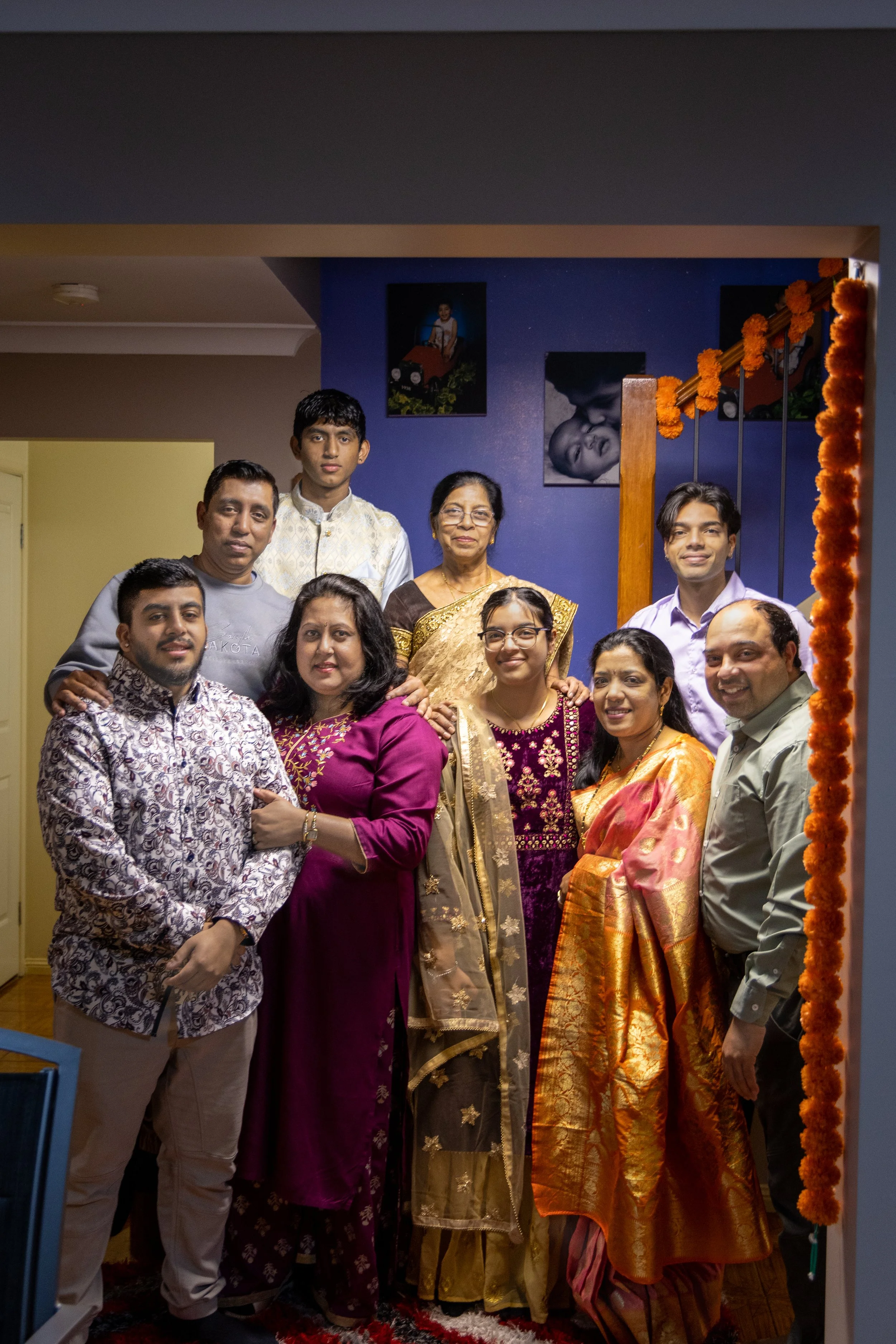 A group of ten people, including men and women, dressed in traditional Indian clothing, posing together in a living room decorated for a celebration with marigold garlands on the staircase. There are family portraits on the blue wall in the backgroun