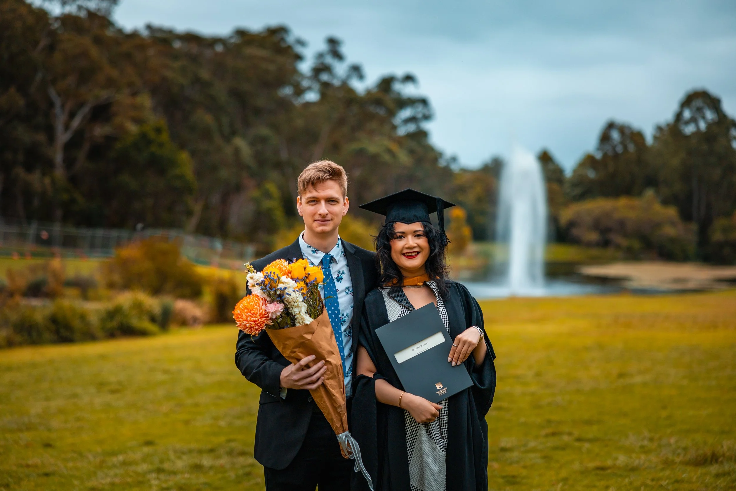 A young woman in a cap and gown holding a diploma, standing next to a young man holding a bouquet of flowers, outdoors in a park with a fountain and trees in the background.