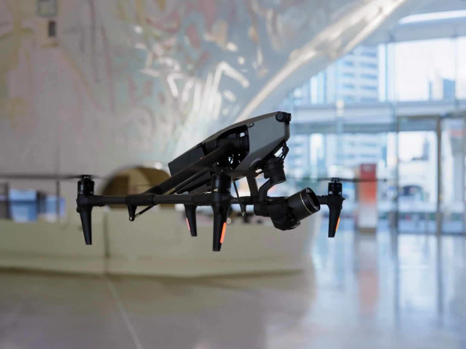 A black quadcopter drone hovers indoors near a window with city buildings in the background.