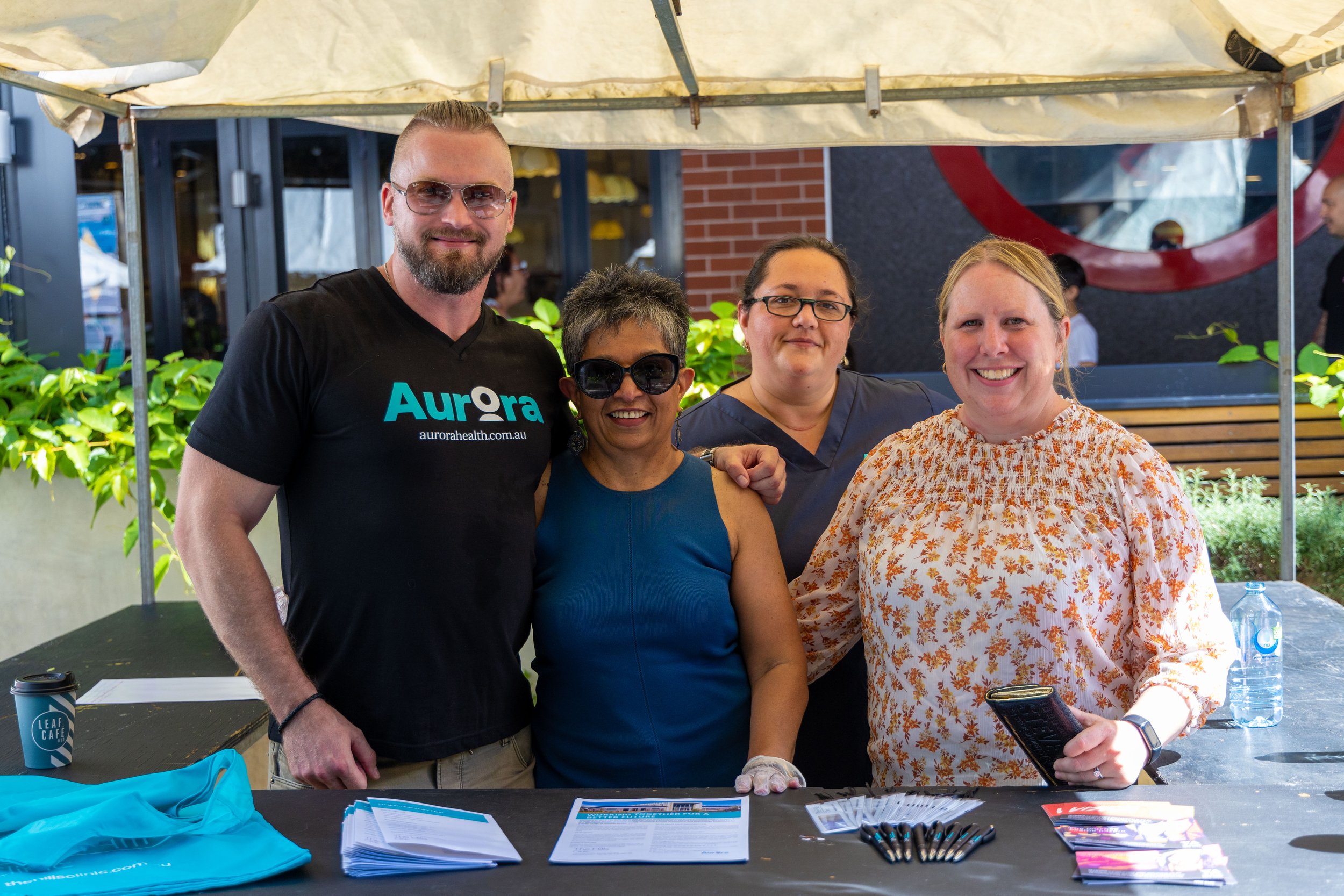 Four people standing behind a table at an outdoor event, smiling at the camera. The table has pamphlets, pens, and a water bottle. The group includes two women and two men, with greenery and a building in the background.