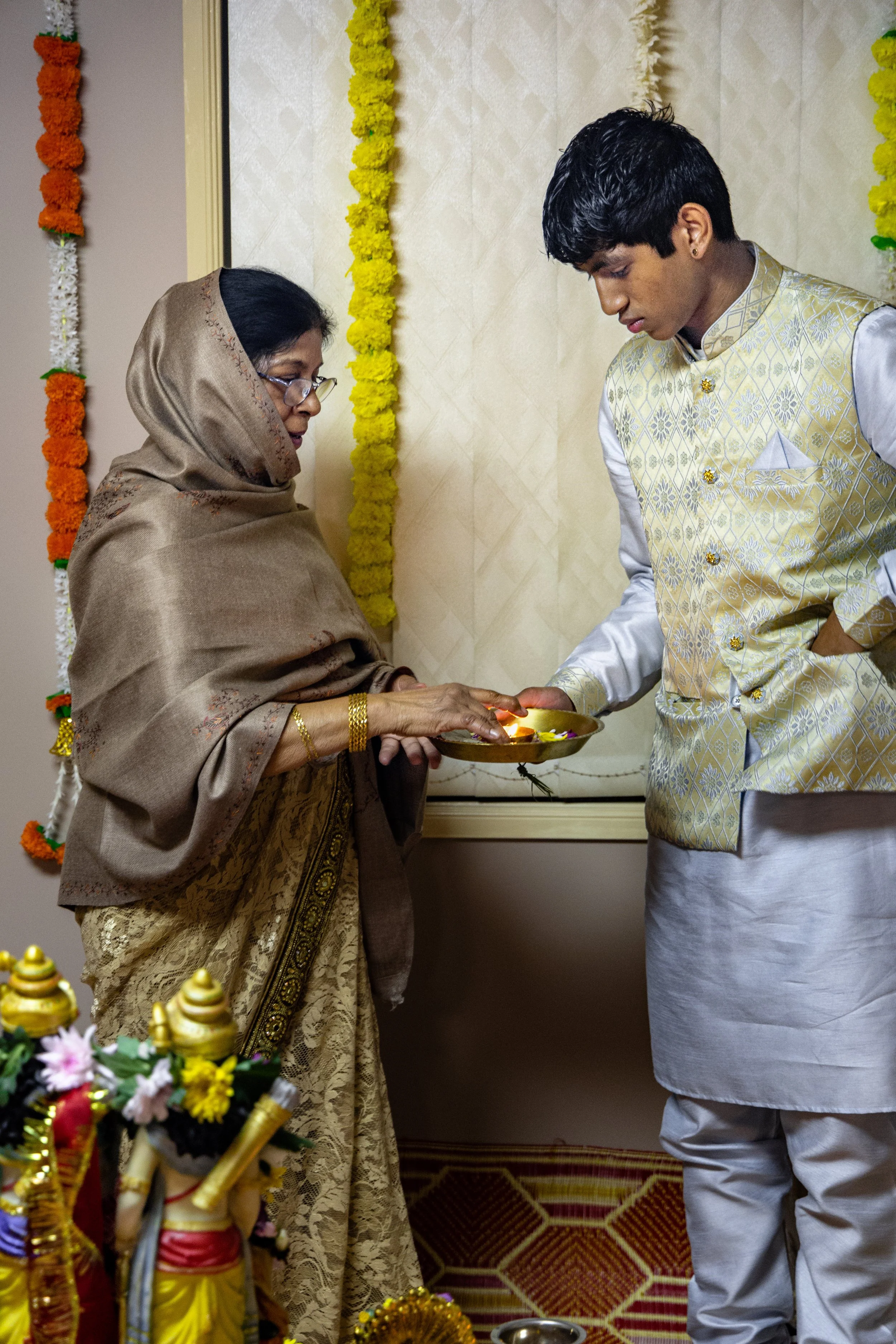 A woman and a young man dressed in traditional Indian attire exchange a lit lamp during a ceremony, with marigold flower decorations in the background.