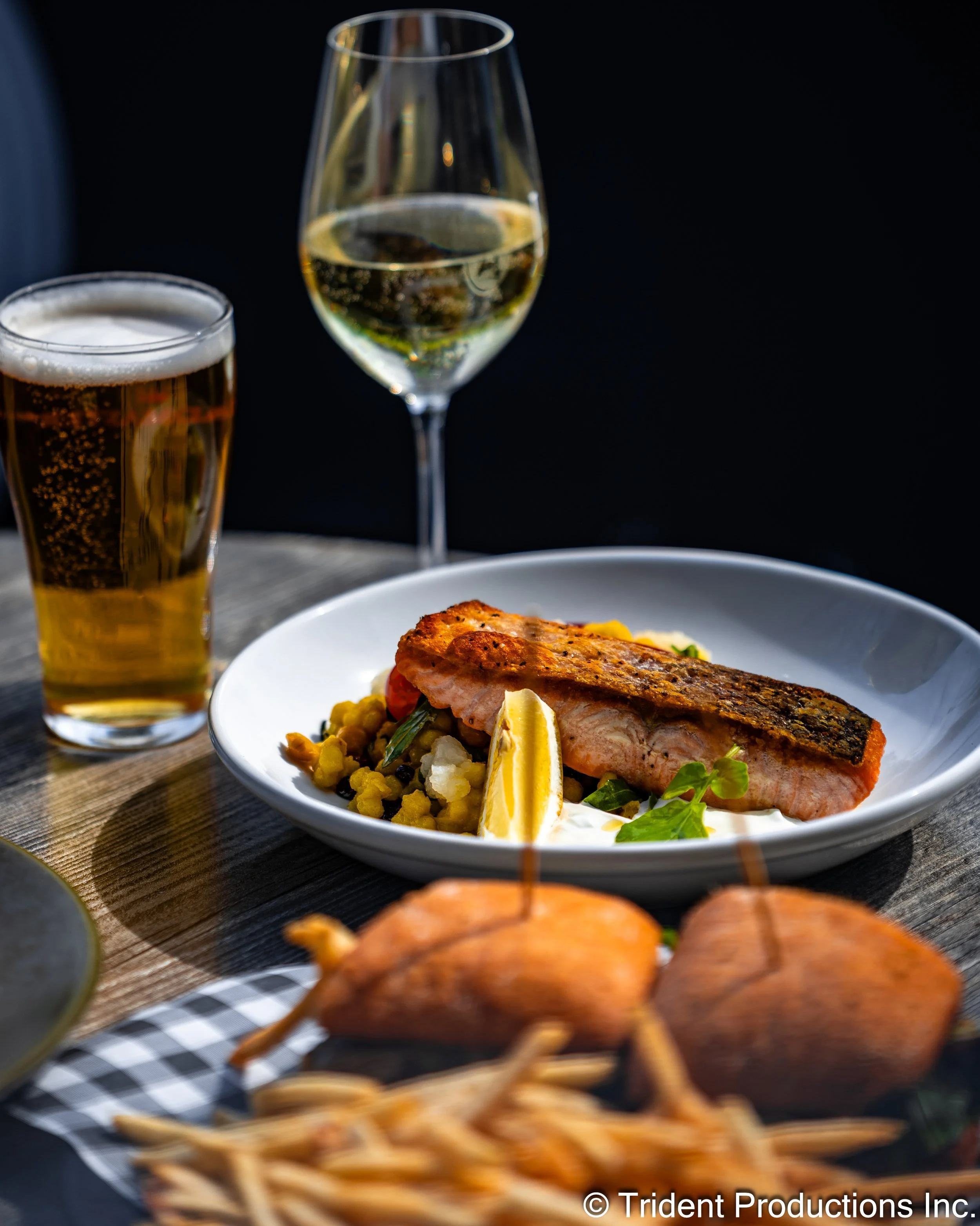 A table with a plate of cooked salmon, lemon wedge, and vegetable side, two drinks (beer and white wine), and a small platter of chicken wings and French fries, set against a dark background.