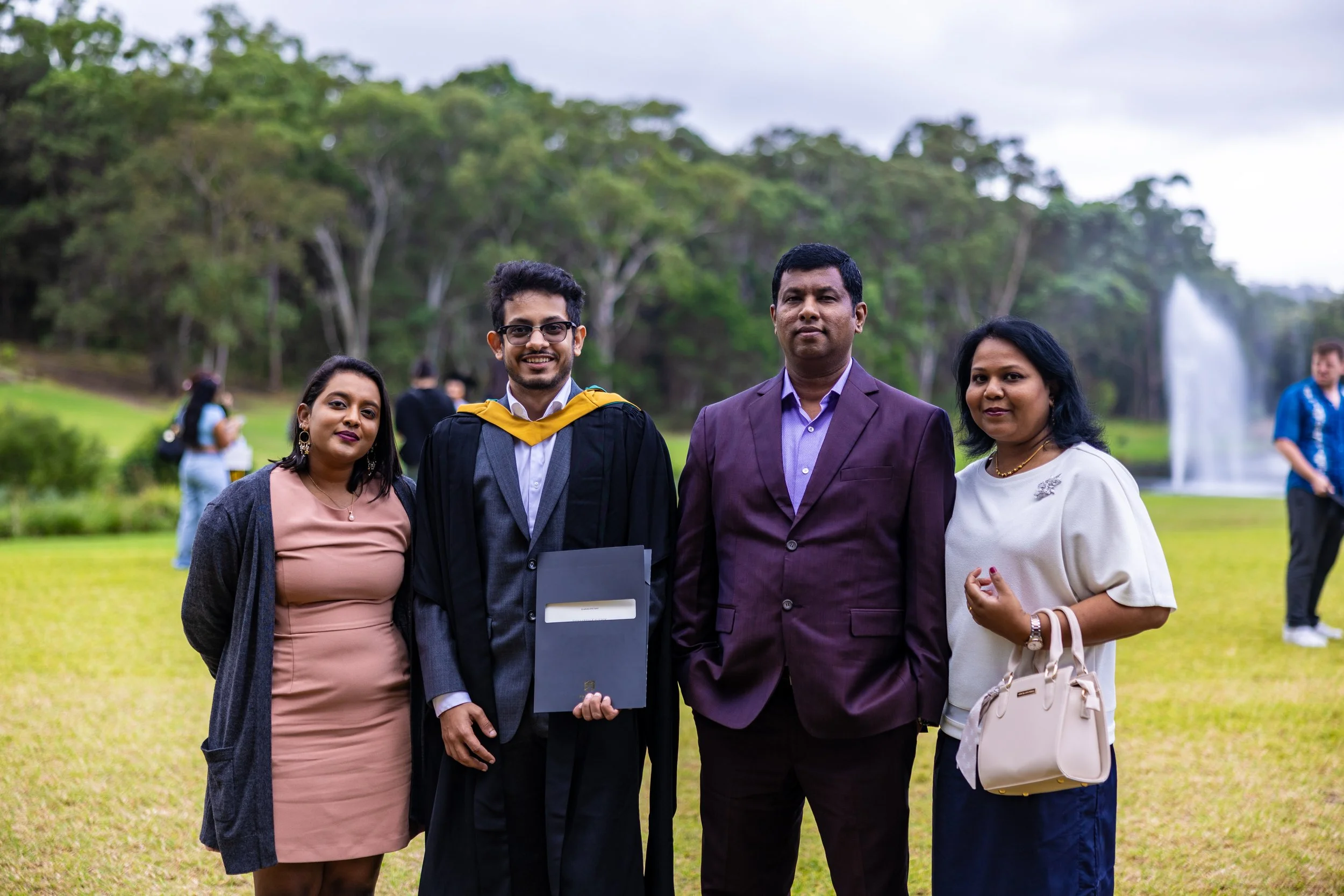 Group of four people standing outdoors celebrating a graduation, with a graduate in cap and gown holding a diploma, in a park with trees and a fountain in the background.