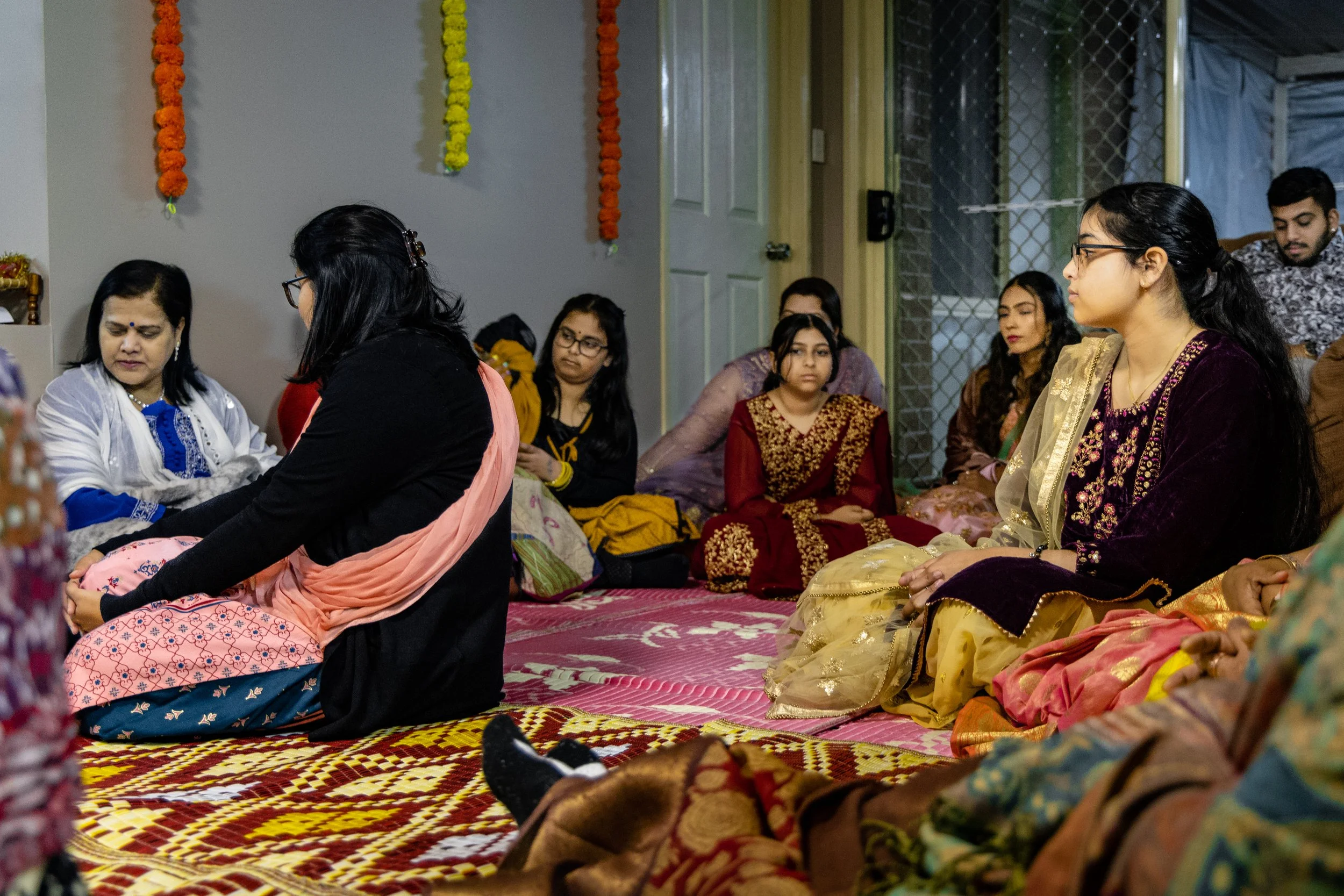 Group of women sitting on the floor wearing traditional Indian attire, with one woman kneeling and appearing to pray or participate in a ceremony, while others observe. The room is decorated with marigold garlands.