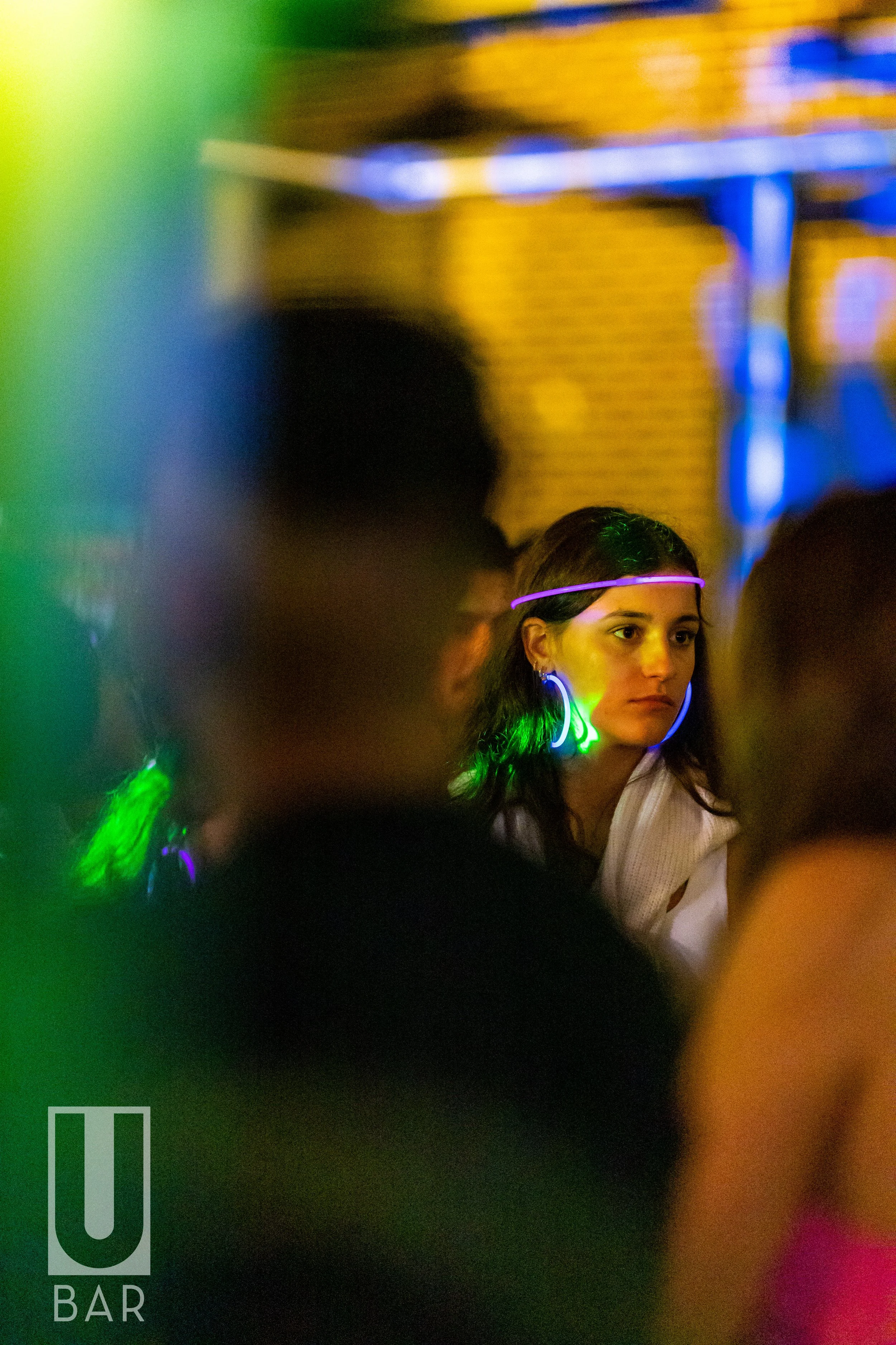 A woman at a bar wearing glow-in-the-dark accessories, including a headband and earrings, surrounded by other people, with colorful lighting.