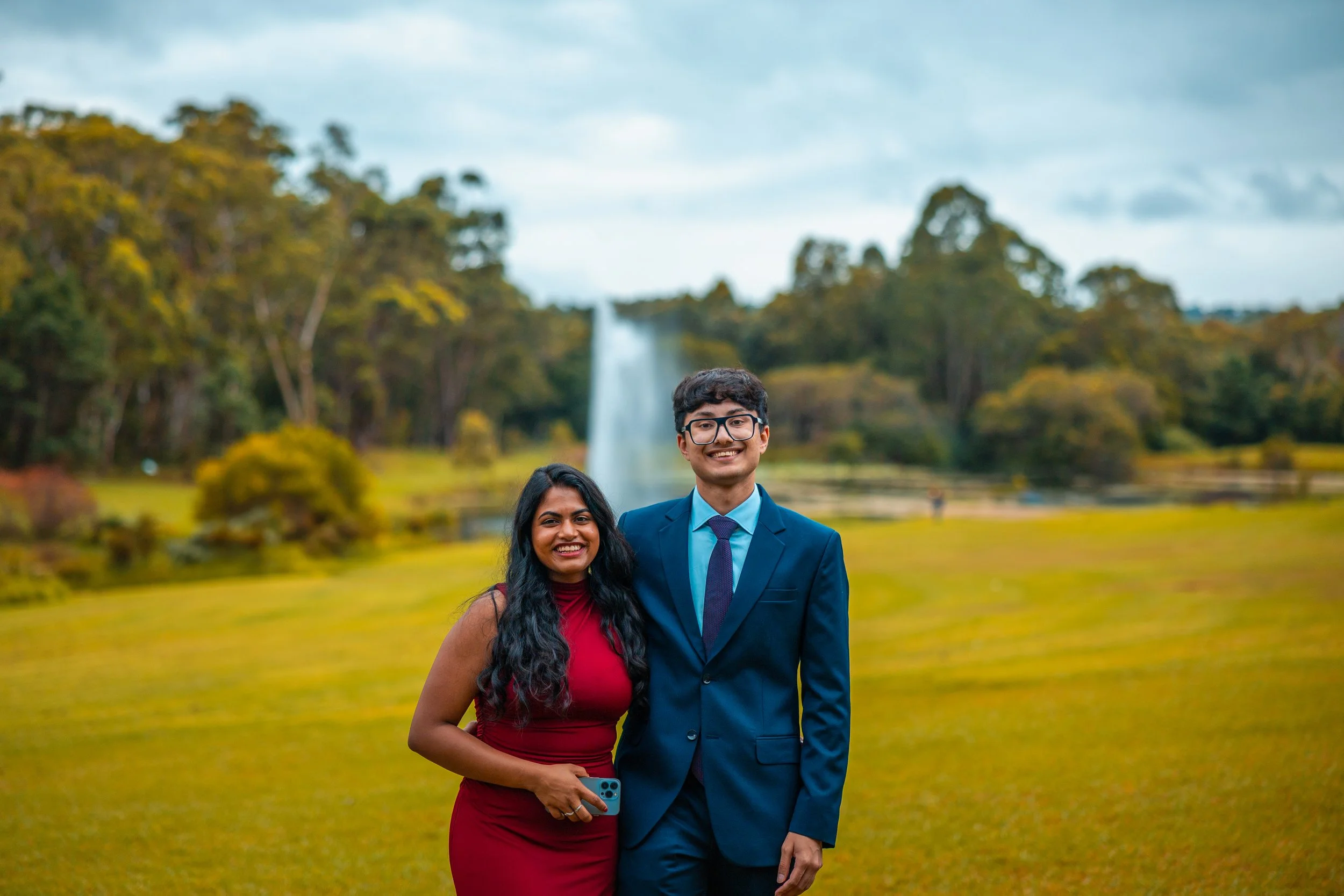 A smiling woman in a red dress holding a phone and a smiling man in a blue suit with a tie, standing outdoors with a lake, trees, and a waterfall in the background.
