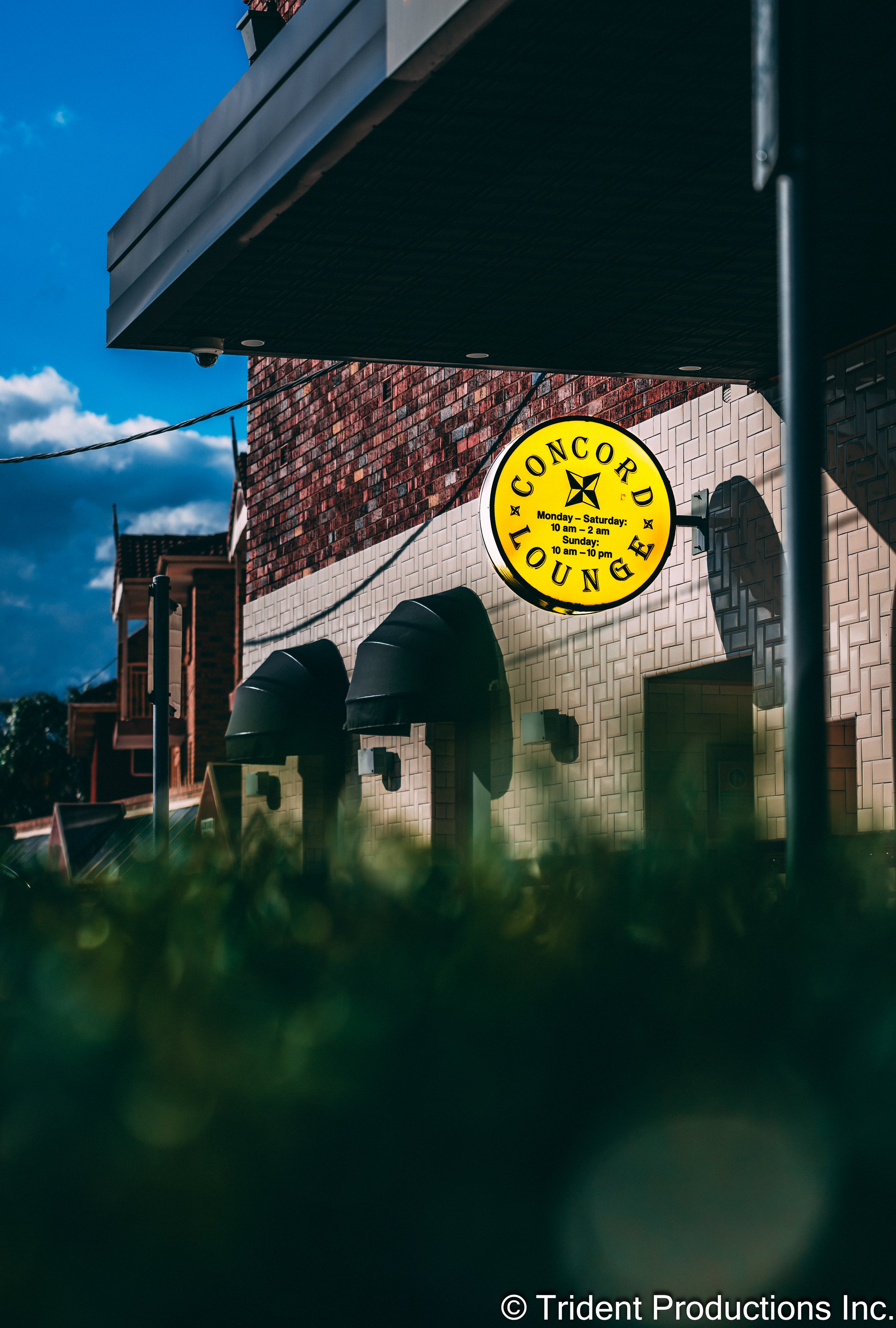A yellow circular sign that reads 'Concord Lounge,' with operating hours and a cross symbol, mounted on a brick building with black awnings and a partial view of a neighboring building, under a partly cloudy sky.