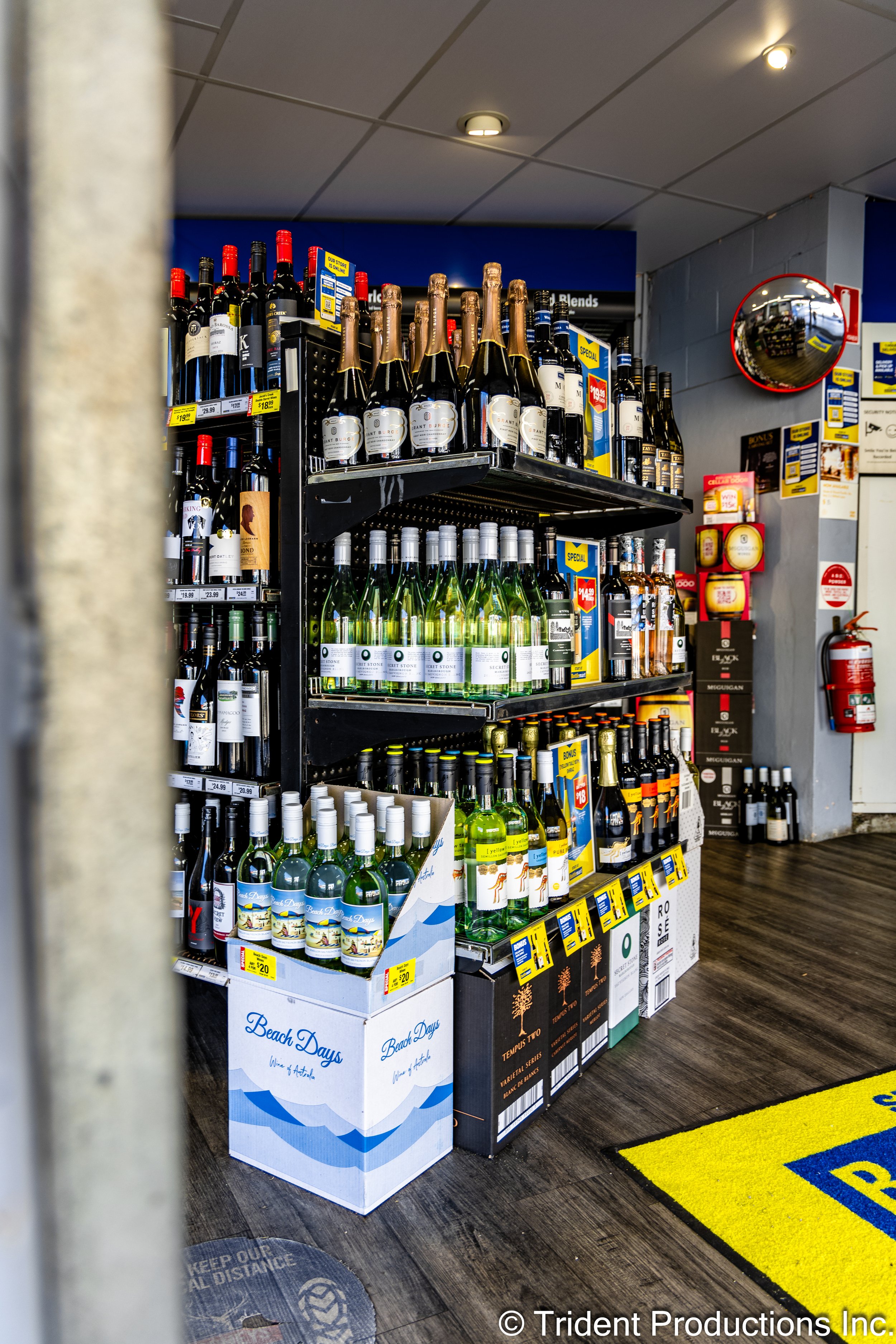 Display of wine bottles on store shelves and in boxes at a retail location, with a fire extinguisher and safety signs visible on the wall.