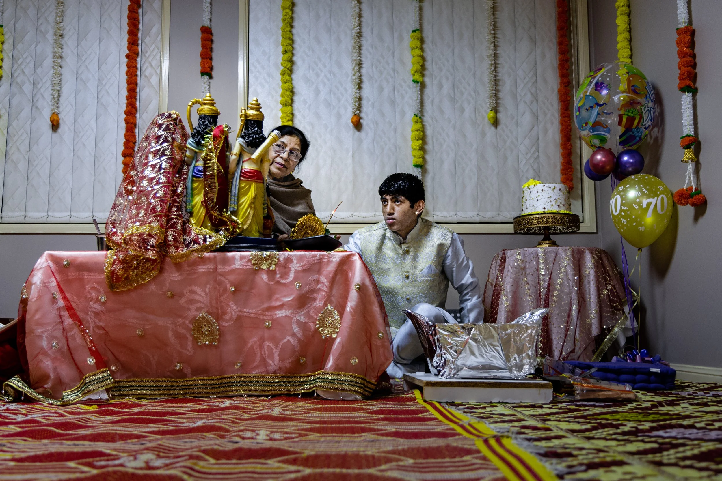 A young man and an older woman at a festive celebration table, with colorful balloons, flowers, and a decorated cake in the background.